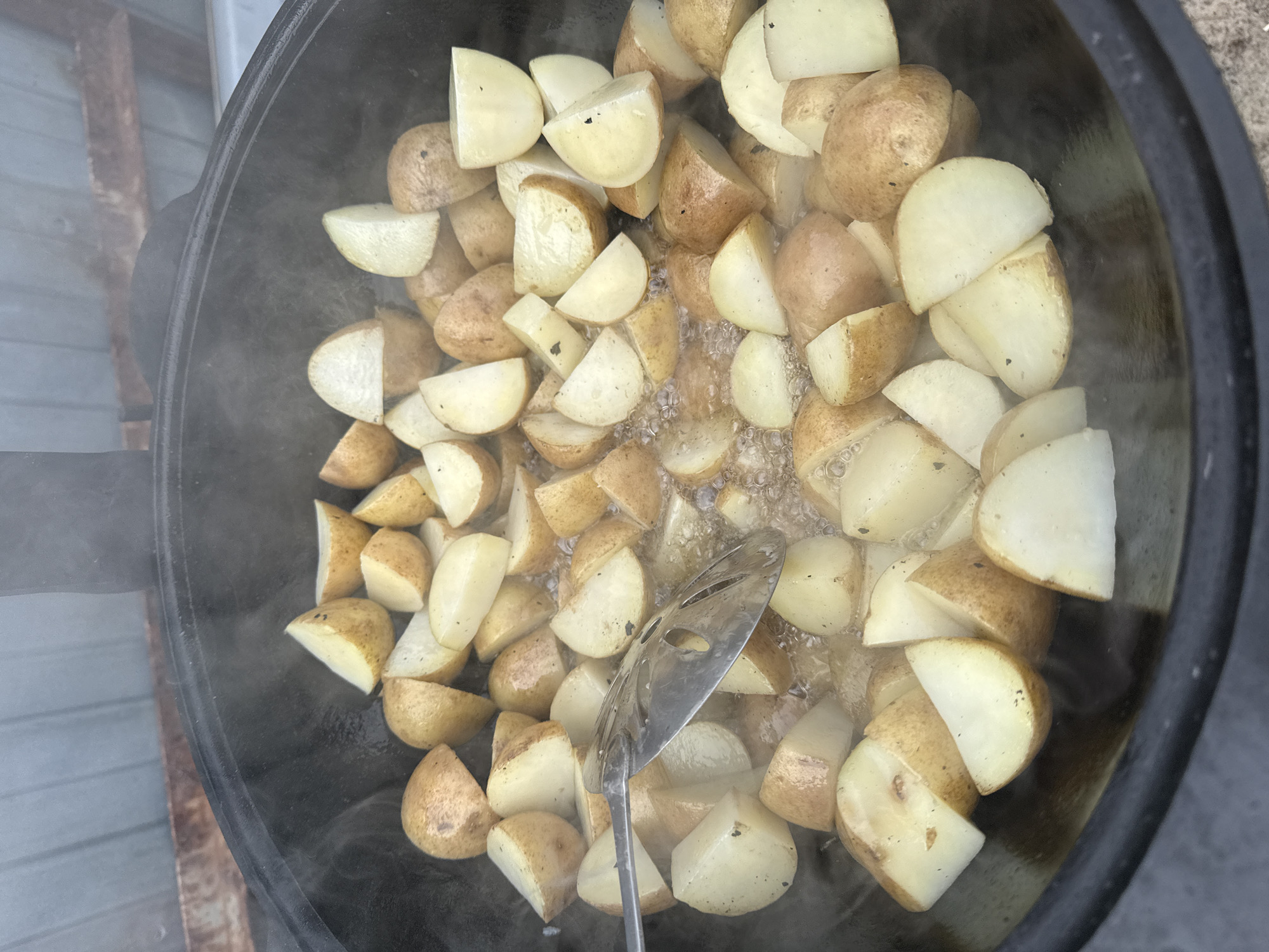 A high-angle shot shows a large black cast-iron pot filled with halved potatoes being cooked in a liquid that appears to be oil or melted fat. A metal spatula is visible within the pot, presumably being used to stir or turn the potatoes. Steam is rising from the pot, indicating the contents are hot. The location is inferred to be in the city of Верхнеказымский, Russia.