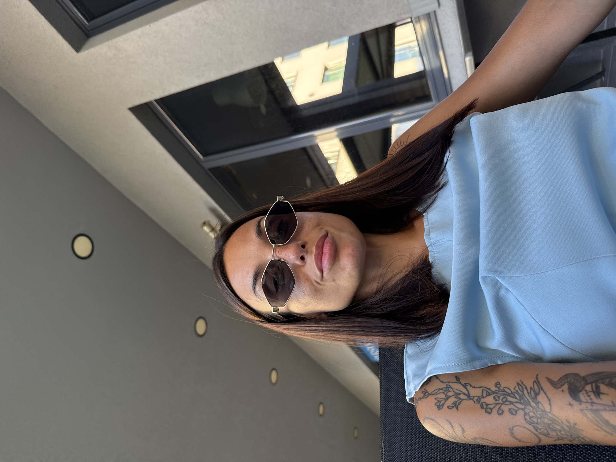 A young woman with dark hair, wearing sunglasses and a light blue top, is seated outdoors, likely on a patio or balcony. She has tattoos on her left arm. Behind her, there is a building with large windows and an overhang with circular lights. The scene is likely a cafe or restaurant in Pleven, Bulgaria, given the architectural style and the typical outdoor seating areas found in the city. The lighting suggests it is daytime, possibly late afternoon or early evening.