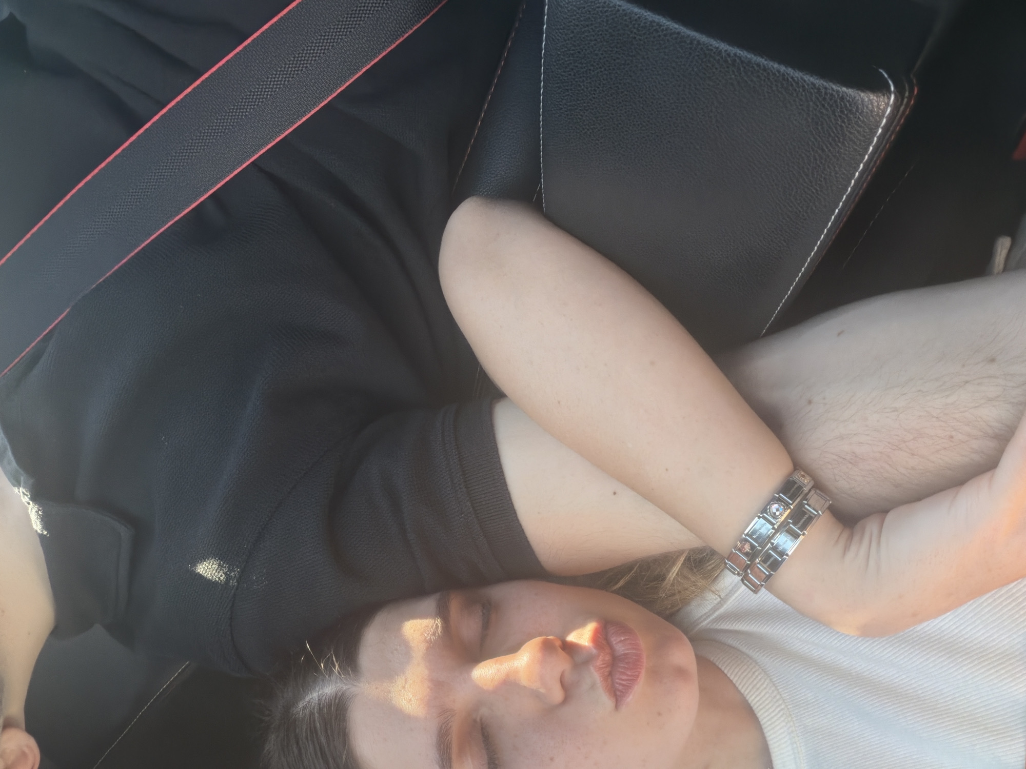 A person is reclining in a car. The person is wearing a black shirt, a white shirt, and a silver watch. They are also wearing a seatbelt. The car has black leather seats. It is not possible to determine if the image was taken in Barakovo, Bulgaria.