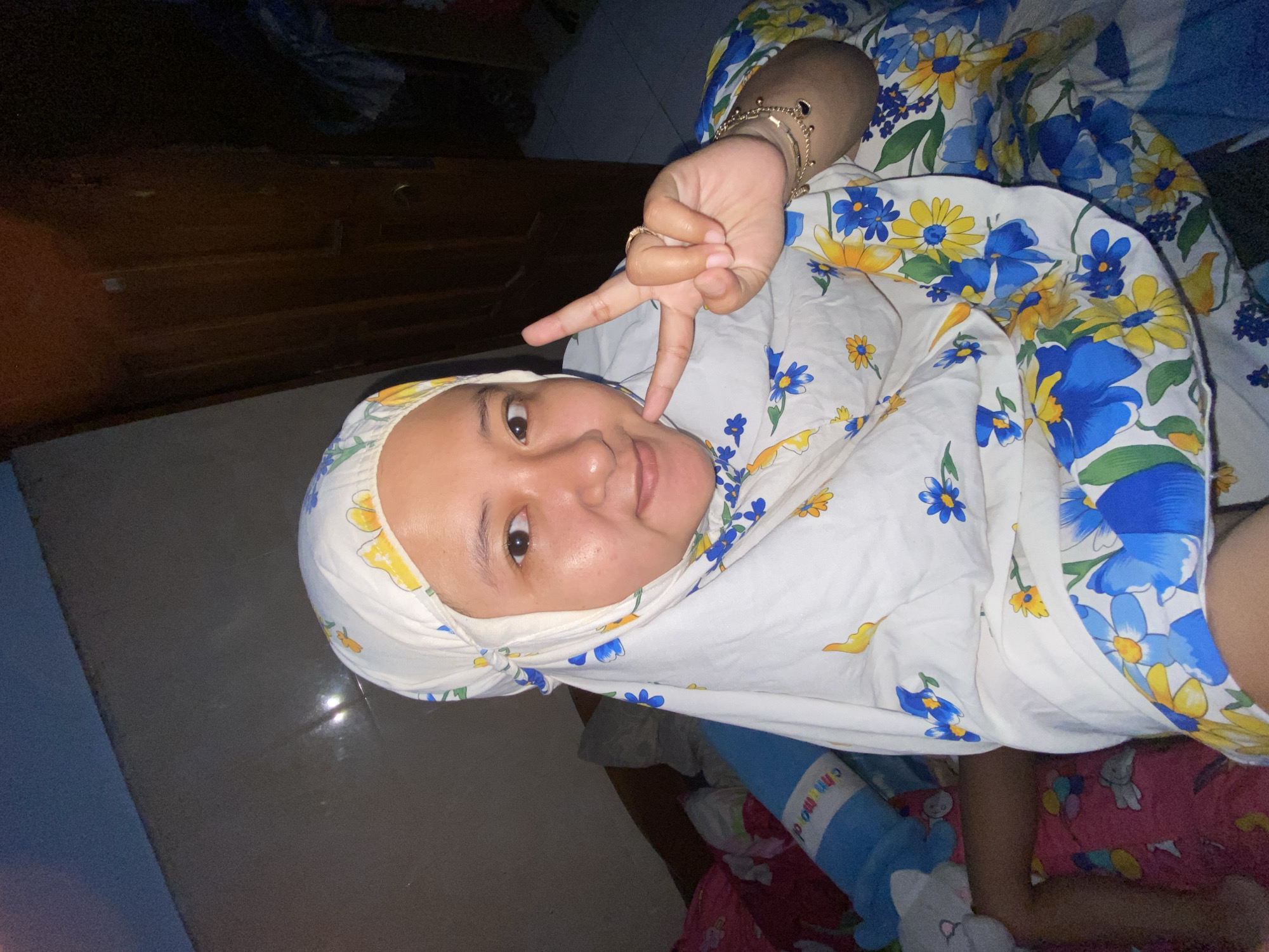 The image is an indoor selfie of a young woman with brown skin. She is wearing a white floral hijab and a floral patterned shirt or blanket with blue and yellow flowers on a white background. She is making a peace sign with her right hand near her face. She is lying on a bed or surface with a pink and red patterned sheet. A blue and white cylindrical object is near her. The background includes a dark wooden cabinet and a partially visible blue wall. The location is likely in Gelam, Indonesia.