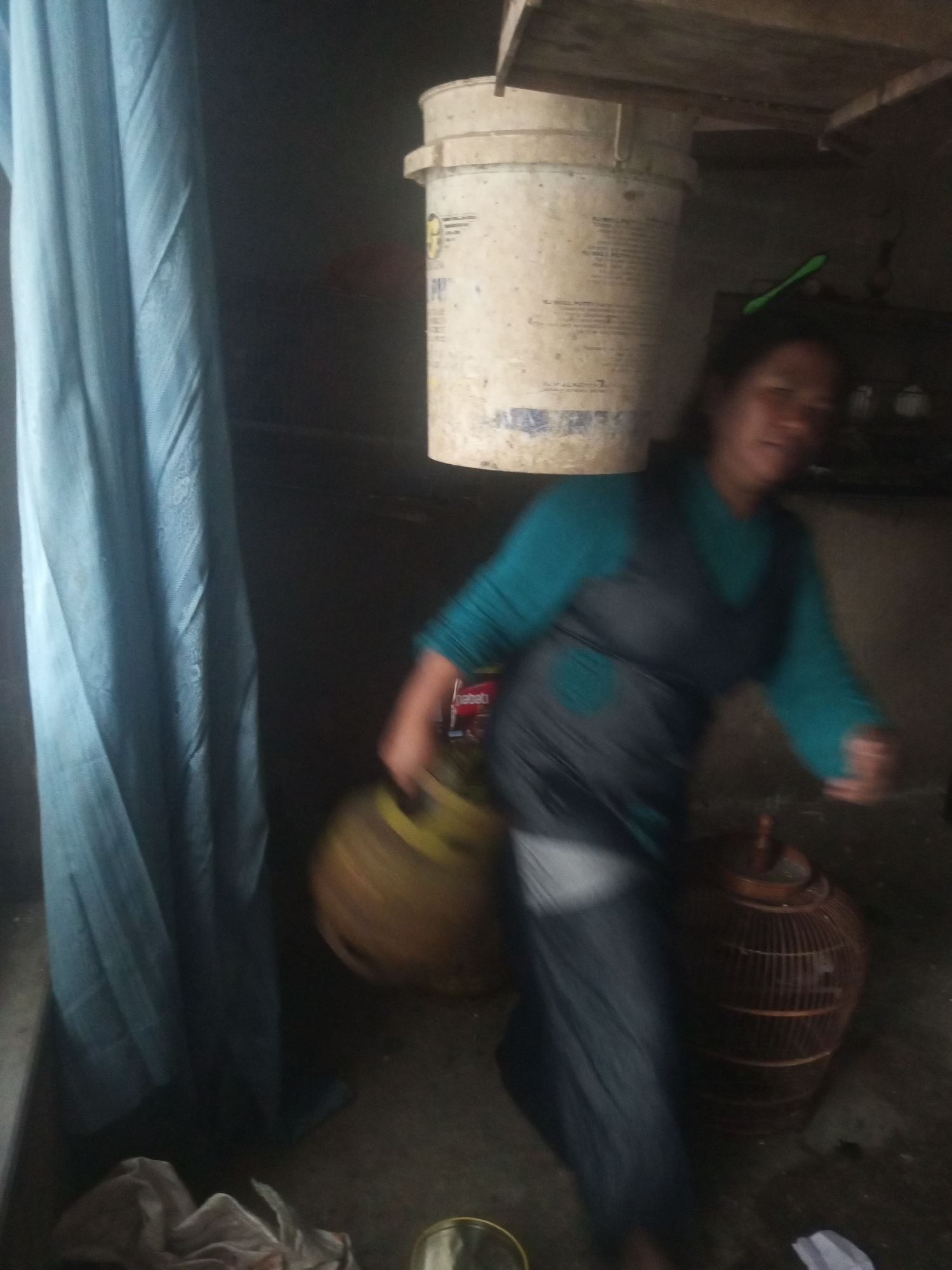 In Andongsili, Indonesia, a medium-shot image shows a person carrying a bucket on their head in what appears to be an indoor or sheltered outdoor space. The person is wearing a teal-colored long-sleeved shirt and a dark gray apron or dress over dark pants. Their body is blurred due to motion. In the foreground to the left, a light blue draped fabric is visible. To the right of the person, a round, brown woven basket is present. The background is dark and indistinct, with a large white bucket perched on a wooden structure above the person's head. A green object, possibly a toothbrush, is positioned on the white bucket. The floor is dirt or concrete.