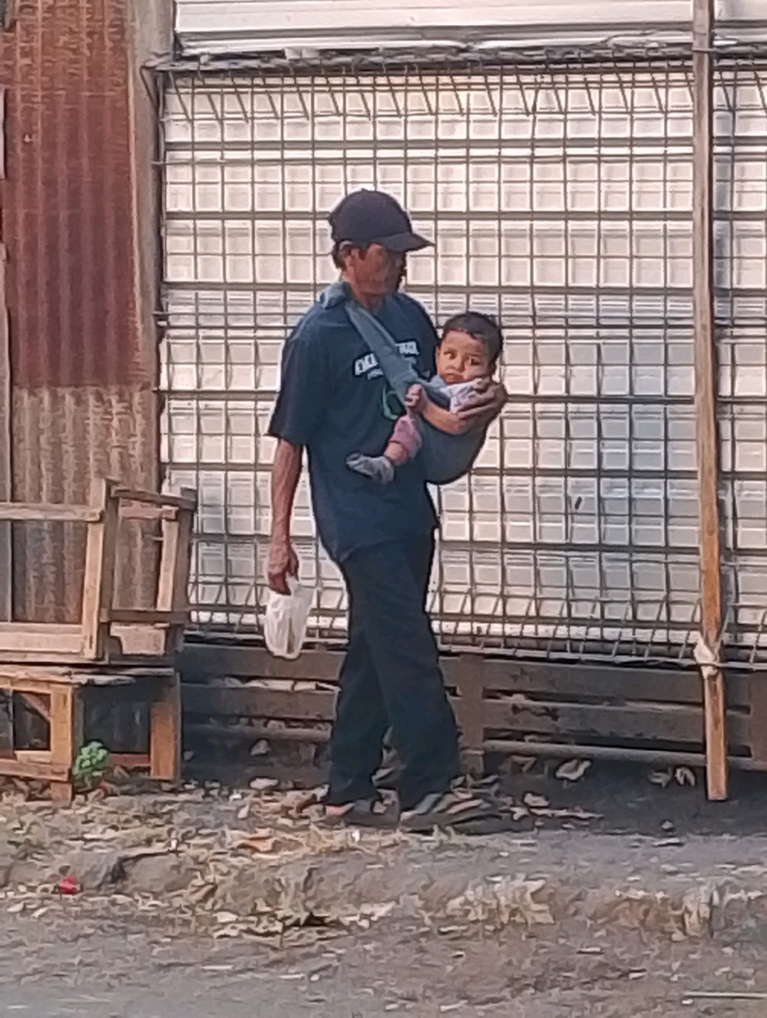 A man wearing a dark baseball cap and a dark t-shirt walks with a baby in a fabric carrier slung over his shoulder. He is carrying a white plastic bag in his right hand. The baby is looking towards the camera. The man is walking on a rough, dirt path with scattered debris. Behind him, there is a corrugated metal wall on the left and a large structure with a grid-like pattern of metal bars and what appears to be translucent panels. In the foreground, there are wooden crates or pallets and more debris on the ground. The scene suggests an outdoor urban or semi-urban environment. This image was taken in Probolinggo, Indonesia.