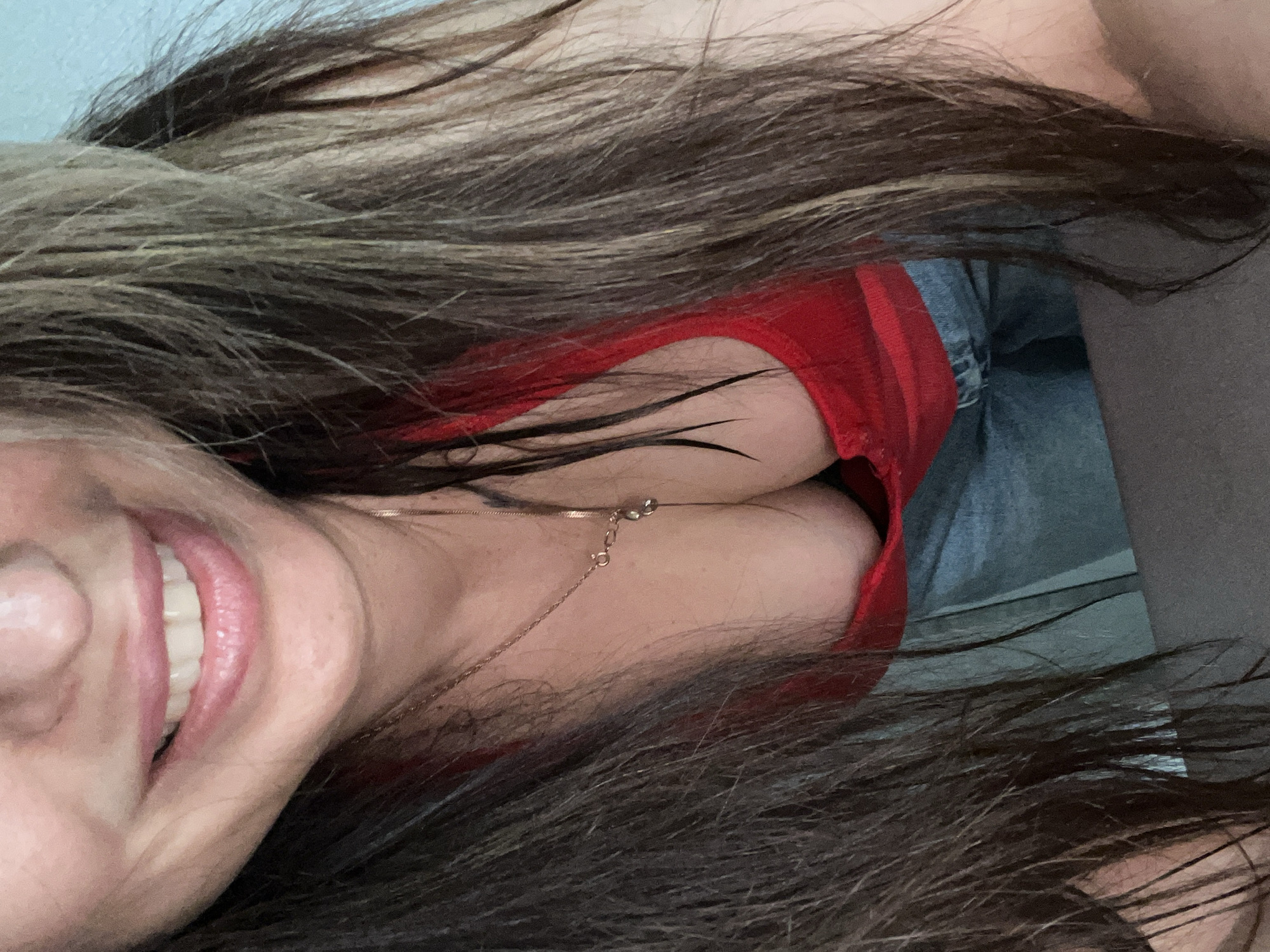 An overhead close-up shot captures a person, likely a woman, looking up at the camera and smiling widely. The image is oriented with the person's head on the left side, angled slightly upwards, and her body extending to the right. Her long, dark brown hair with lighter, possibly blonde or light brown, highlights cascades over much of the frame, creating a sense of intimacy.
The subject's face is partially visible on the far left, dominated by a radiant, open-mouthed smile that reveals white teeth and pink lips. A delicate gold-colored chain necklace is visible around her neck and collarbone. She is wearing a bright red top, possibly a tank top or camisole, and glimpses of light blue denim clothing, likely jeans or shorts, are seen further down. Parts of her bare skin are visible beneath her hair and clothing.
The background is indistinct, showing light-colored surfaces in the upper left corner and darker tones in the lower right, suggesting an indoor setting. The lighting appears soft and even. The overall impression is one of happiness and casual intimacy. No text is visible in the image.