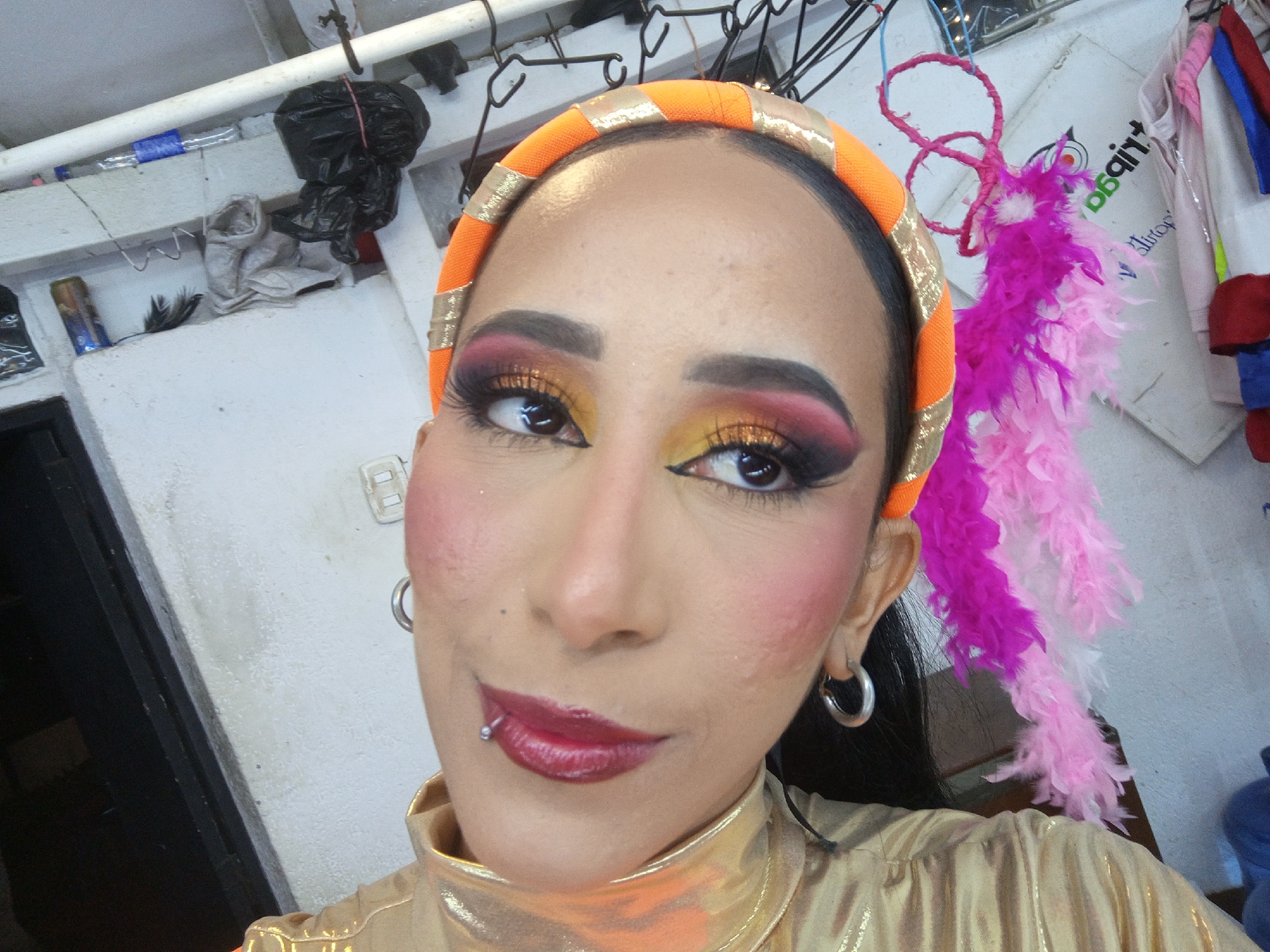 A person with dark hair, heavy makeup, an orange and gold headband, and a gold shirt poses for a selfie. They have pierced ears with hoop earrings, and a lip piercing. A pink and purple feather boa is visible on one side of their head. The background includes white walls, a door frame, miscellaneous items on a shelf, and clothing hanging from hooks. The location is likely indoors. There is no verifiable evidence within the image to confirm that it was taken in the city of Porlamar, Venezuela.