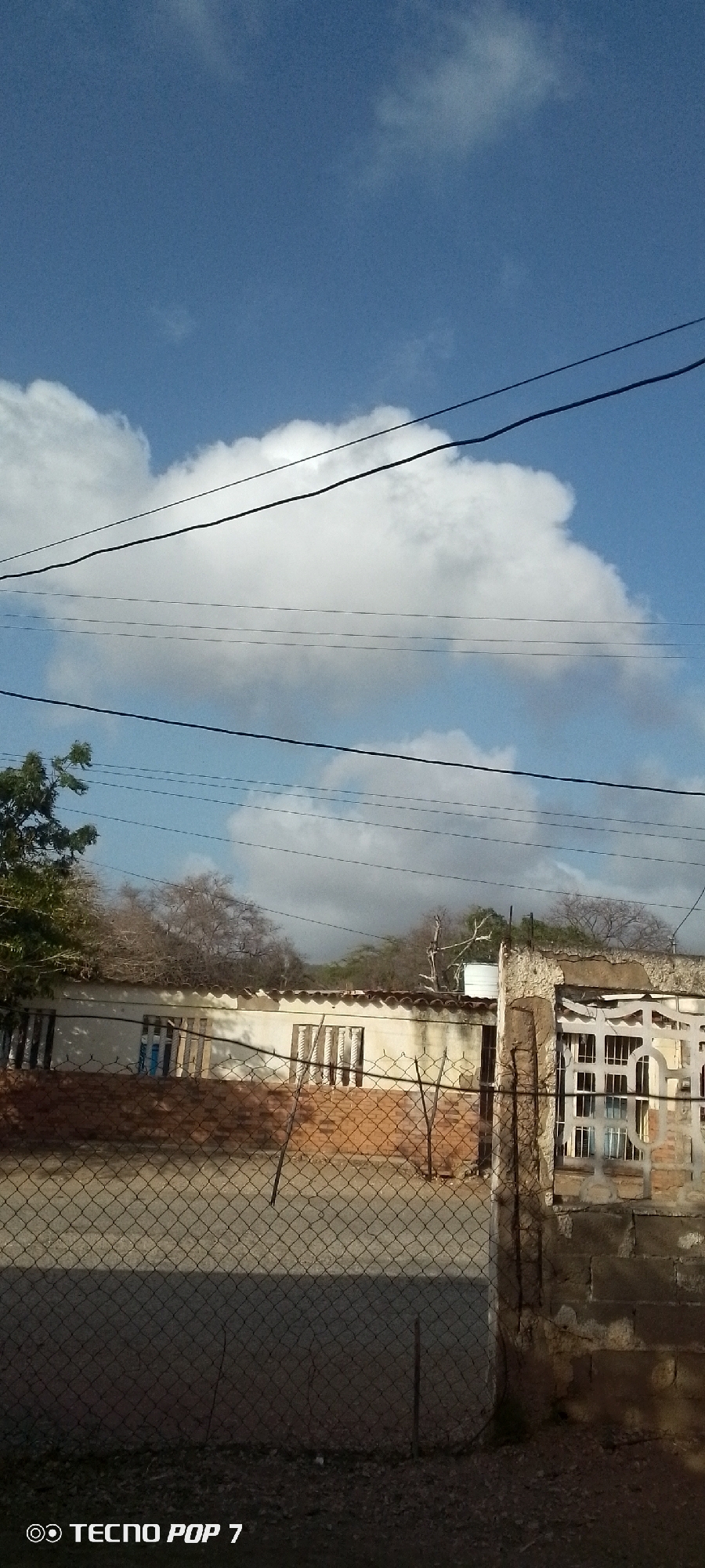 The image shows a building in Altagracia, Venezuela under a partly cloudy sky. Electrical wires cross the frame in the sky. A chain-link fence runs in the foreground. The building behind the fence has a damaged wall section to the right, adjacent to a window. The walls of the building are painted white with red brick visible below the windows. Some trees are visible behind the building. The Tecno Pop 7 label is visible in the lower left corner.