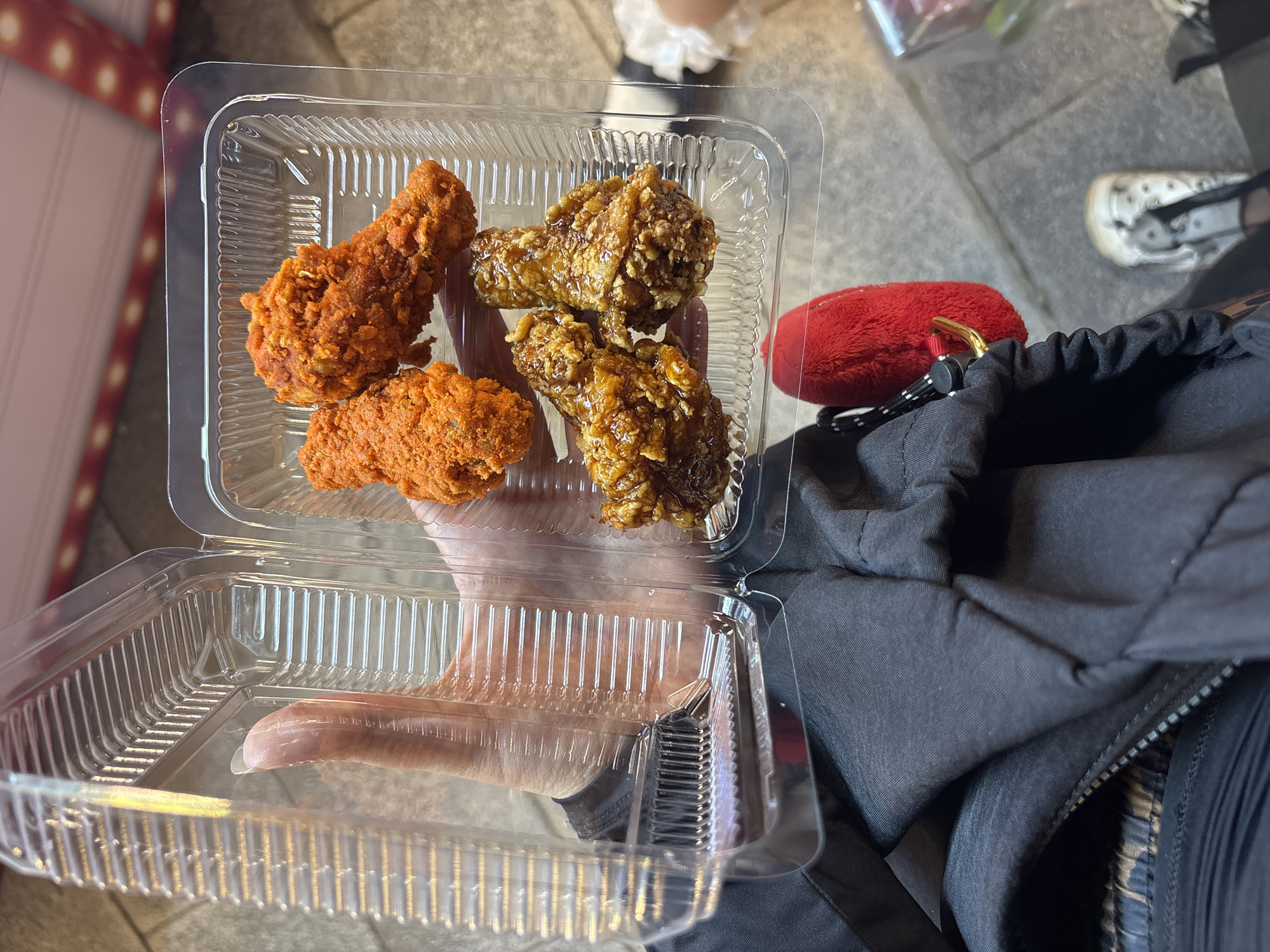 A person holds an open, clear plastic container filled with fried chicken. The container is divided into two compartments. The left compartment is empty and lies open towards the viewer. The right compartment contains five pieces of fried chicken. Two pieces are coated in a bright orange, crispy batter. Three pieces are coated in a darker, glazed sauce that appears sticky. The person's hand is visible, holding the open lid of the container. They are wearing a dark jacket with a zipper. A small, red, fuzzy keychain is attached to the jacket. The background appears to be an outdoor urban environment, with cobblestone-like paving stones visible.
