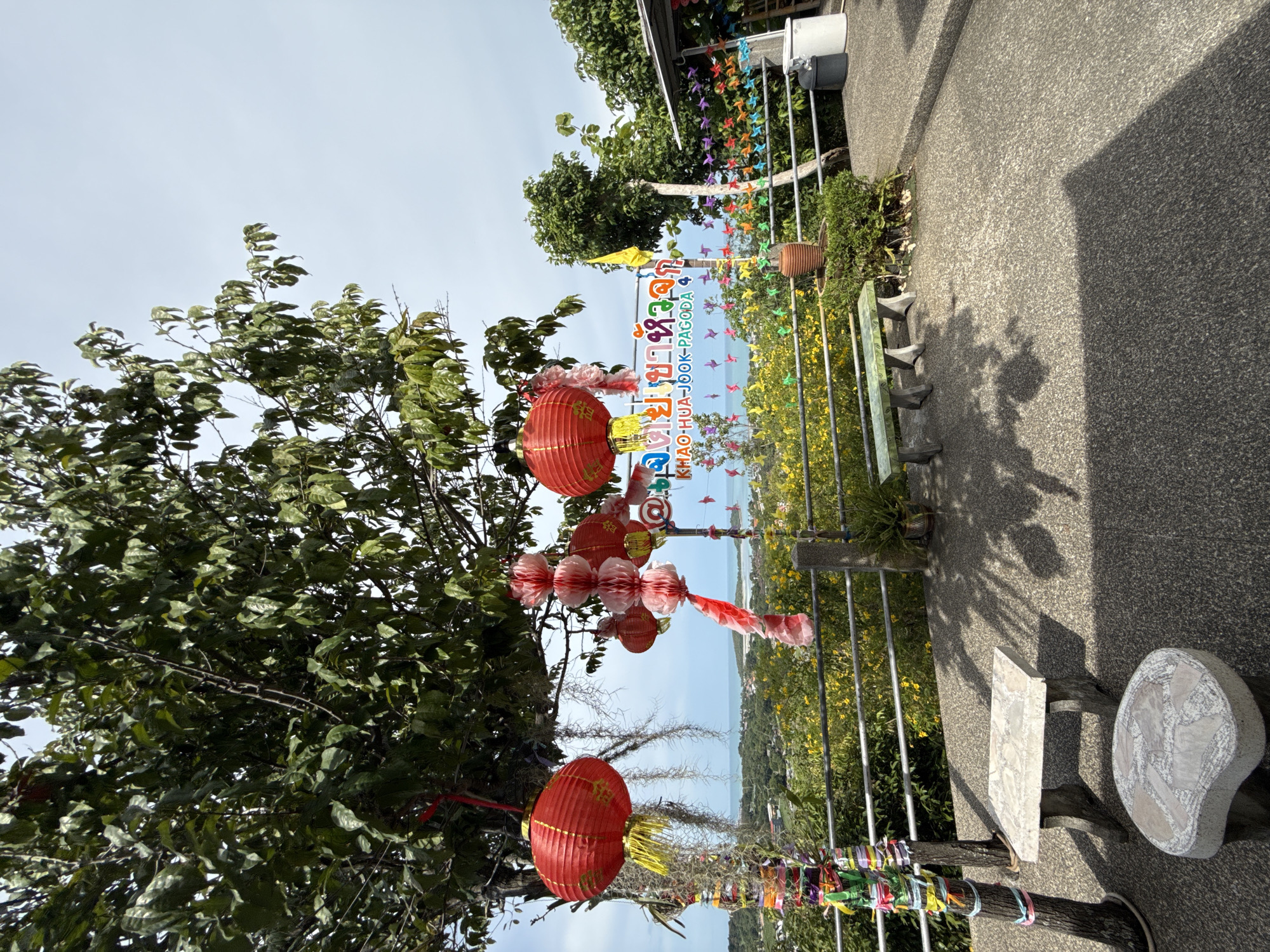 A wide-angle, eye-level shot captures an outdoor seating area at a scenic overlook in Ban Bang Rak, Thailand. The foreground is dominated by a grey, speckled concrete pathway and two stone benches, one with a rectangular top and the other a rounded seat. To the left, a tree trunk is wrapped with colorful ribbons. Red Chinese lanterns hang from the branches of a large tree in the mid-ground, along with decorative pink paper lanterns and streamers. Behind the seating area, a metal railing separates the pathway from a hillside covered in yellow flowers and lush green foliage. Further in the background, the ocean is visible with a distant coastline dotted with buildings. A colorful banner with Thai text and "KHAO-HUA-JOOK-PAGODA 4" is mounted on a wooden pole. Various colorful paper pinwheels are strung along the railing. A yellow flag flies from another pole. A potted plant sits on a pedestal, and a large terracotta pot is nestled among bushes. To the right, a small white building with a dark roof is partially visible, along with a dark grey trash can and a large white bin. The sky is overcast with a pale blue hue.