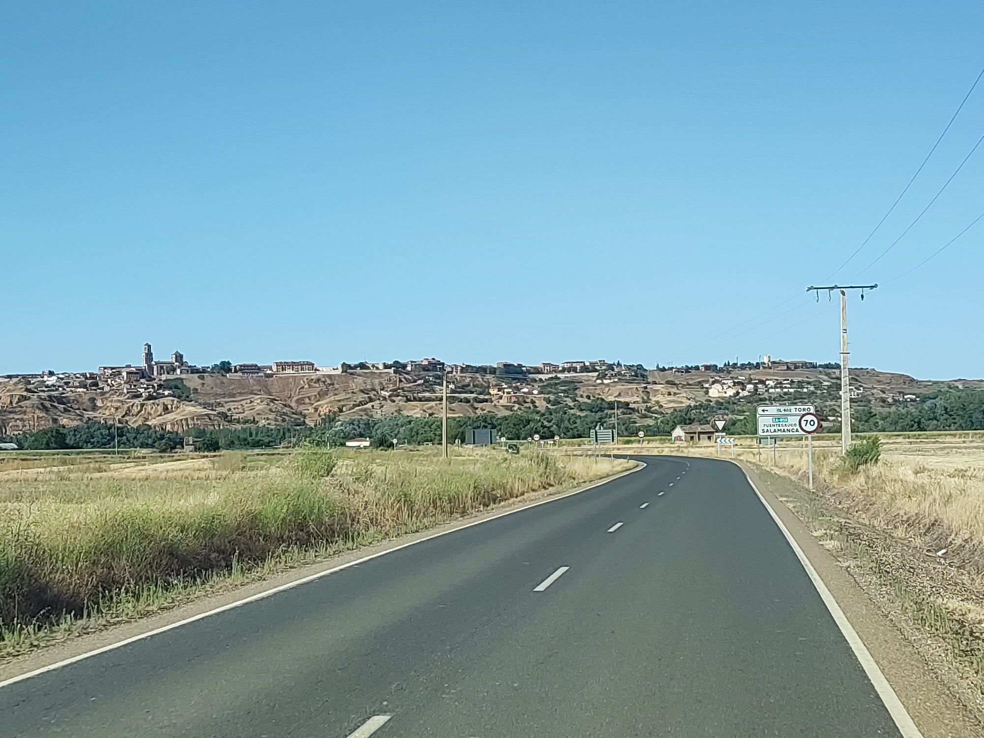 This is a photograph taken from a vehicle traveling on a paved road. The road curves to the left in the foreground, with dry, yellowish grass lining the right side and a mixture of green and dry grass on the left. In the midground, a town is visible on a hillside. The town features buildings with red tile roofs and some taller structures that appear to be churches or towers. The hillside is characterized by eroded earth and sparse vegetation. A utility pole with three wires runs along the right side of the road, extending into the background. A road sign is also visible on the right, indicating "CL-602 TORO" with an arrow pointing left, and "FUENTE<SAUCO SALAMANCA" with a speed limit sign of "70". The sky is clear and blue.