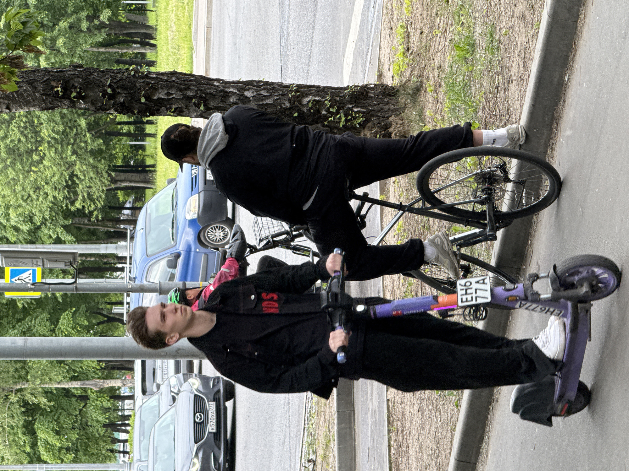 Two individuals are visible on a paved path next to a road in what appears to be an urban or suburban setting in Moscow, Russia. One person, wearing a black hoodie with a gray hood and black pants, is on a black bicycle, with their right foot on the ground near the curb. A black baseball cap is worn backwards. The second person is on a purple electric scooter, holding the handlebars. They wear a black jacket over a black shirt with red text, possibly reading "ANDS", black pants, and white sneakers.
The path is bordered by a curb, beyond which is a road with vehicular traffic. A blue car, resembling a van, is parked or moving behind the cyclist. Further down the road, a dark gray Mazda SUV (license plate K570XA 777) is visible. In the background, numerous green trees line the street, suggesting spring or summer. A pedestrian crossing sign (blue square with white walking figure) is also visible. The electric scooter has the identification "EH6 77A" and "EH677A" printed on its stem and side.
The scene is lit by natural daylight, likely overcast given the soft shadows. The individuals appear to be either stopped or moving slowly, possibly waiting or taking a break from their respective rides.