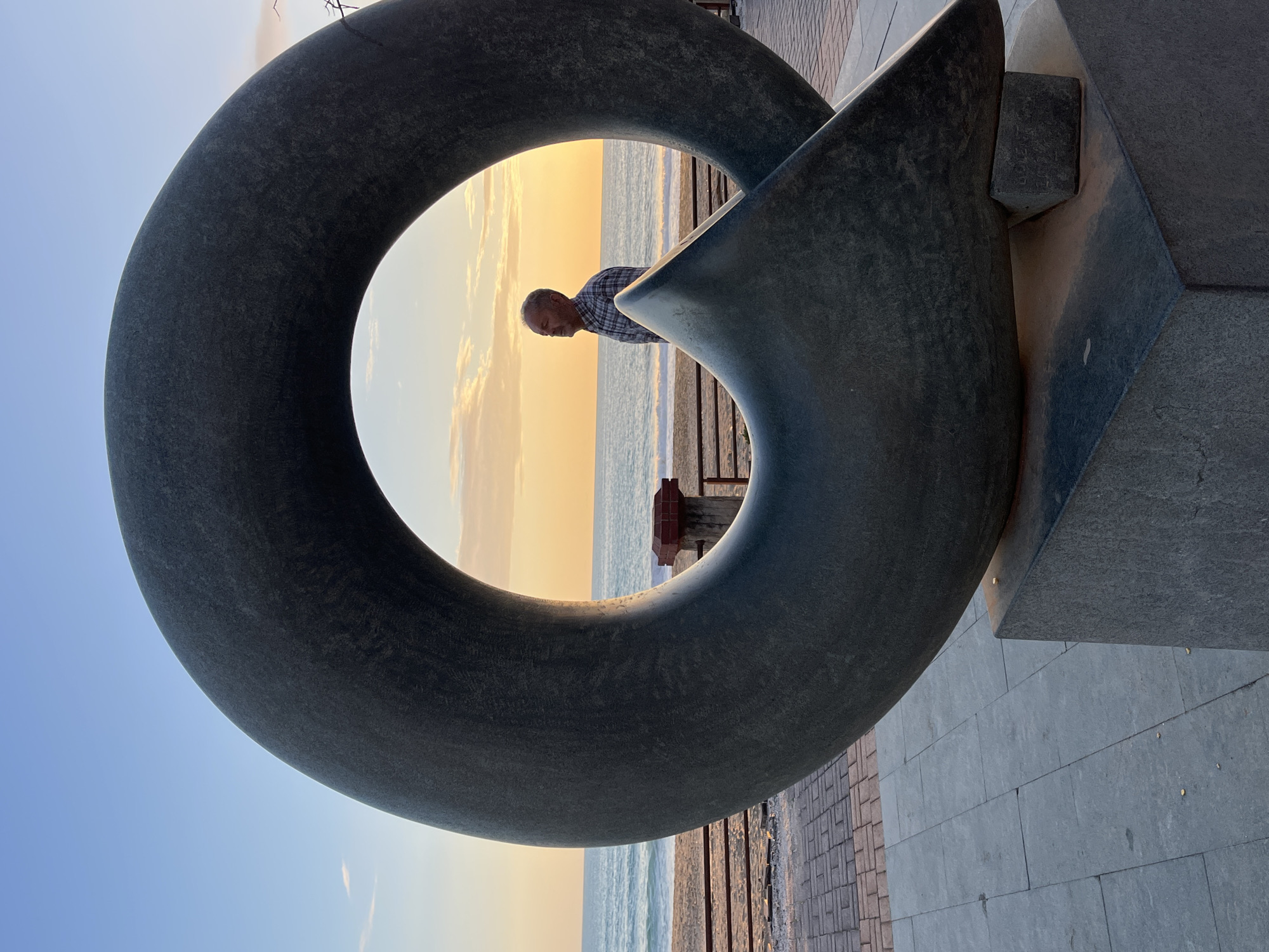 The photograph shows a grey stone sculpture with a large circular opening, positioned on a tiled walkway in Alanya, Turkey. A man wearing a plaid shirt can be seen through the circular opening of the sculpture, sitting on a bench overlooking the beach and the sea, which is illuminated by the sunset. The sky shows a gradient of blue to yellow due to the setting sun.