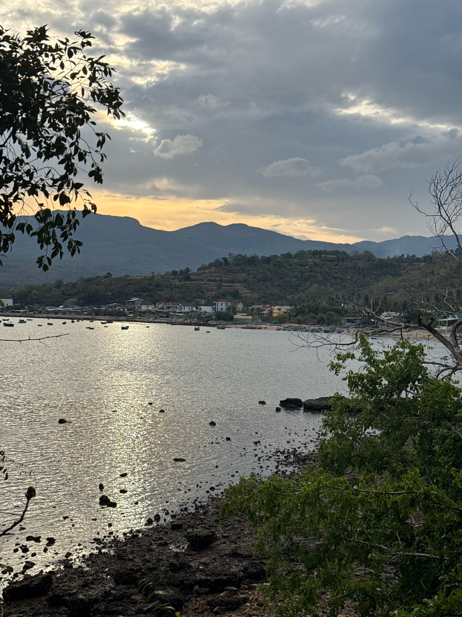 The image depicts a scenic view of the coastline in Song Cau, Vietnam, likely at sunset given the lighting conditions. The foreground features a rocky shoreline with green foliage from trees framing the right and left sides of the image. The calm sea stretches out to meet a distant shore lined with a small village, characterized by houses with red and yellow roofs. Beyond the village, lush green hills rise, culminating in mountainous terrain under a dramatic, cloud-filled sky. Several small boats are visible on the water near the shore, suggesting a fishing community. The sun's rays break through the clouds, creating a shimmering reflection on the water's surface.
