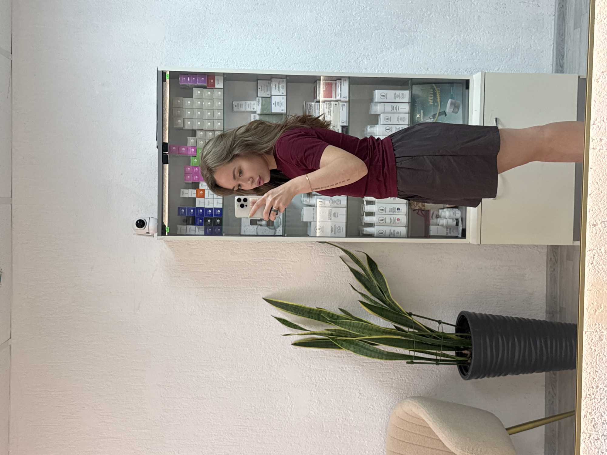 A young woman with shoulder-length brown hair is taking a mirror selfie in what appears to be a salon or beauty shop. She is wearing a dark red t-shirt and dark grey shorts. She is holding a white smartphone in her right hand, angled to capture her reflection. Behind her, a tall glass display cabinet is filled with rows of white boxes and bottles, likely skincare or cosmetic products. To her left, a tall potted snake plant with long, green and yellow striped leaves stands in a black textured pot. A portion of a beige armchair with gold legs is visible in the far left of the frame. The wall behind the plant and the cabinet has a textured white finish. The image appears to be taken in Chaikovsky, Russia.