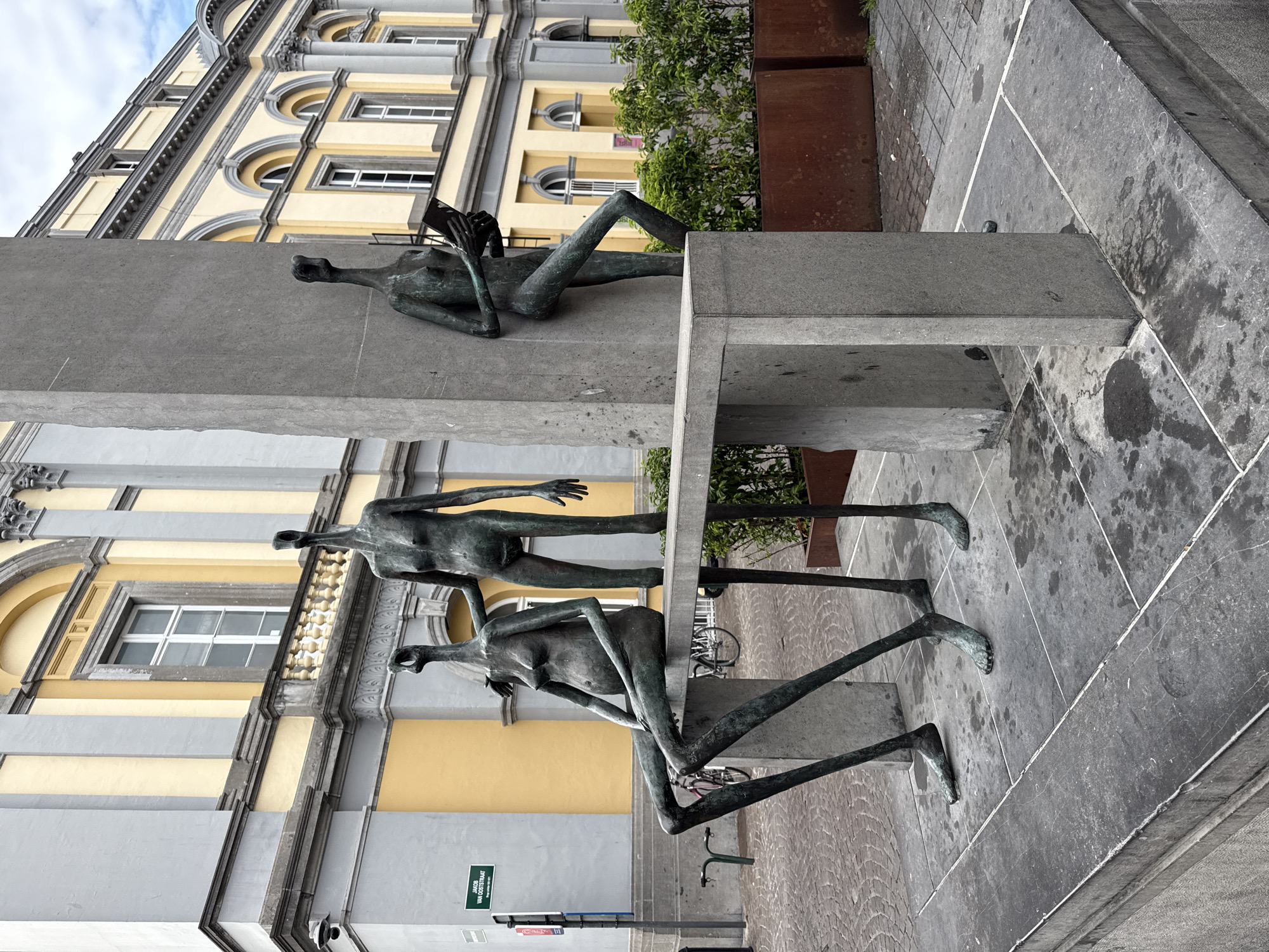 A bronze sculpture of three elongated, stylized human figures is situated in front of a building with a yellow facade and grey accents in Bruges, Belgium. One figure sits on a grey stone bench, leaning back with its legs extended. Another figure stands behind it, with one hand resting on the seated figure's shoulder. The third figure, also standing, is engaged with a book held in its hands. The figures are abstract, with smooth, minimalist forms and indistinct facial features. The bench and a supporting pillar are made of rough-hewn grey stone. Behind the sculptures, a street is visible with cobblestones and a bicycle rack. The building in the background features arched windows and classical architectural elements. Greenery in planters is also present.