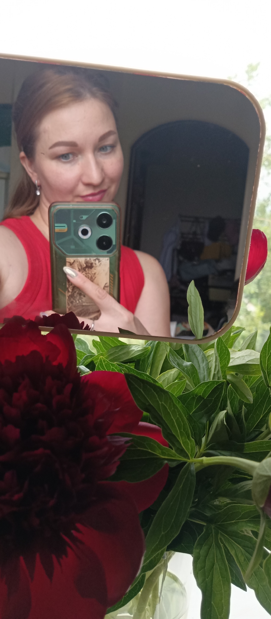 A mirror selfie shows a woman in a red tank top holding a smartphone with a green case and taking a photo of her reflection. She has light skin, blue eyes, and auburn hair pulled back. Her left ear is visible with a dangling earring. The phone's camera module, featuring multiple lenses, is prominent. Behind her in the mirror, a blurred room is visible, containing a person wearing a yellow shirt and possibly some clothes hanging. In the foreground, a dark red peony flower and green leaves fill the bottom portion of the frame. It is impossible to infer the location of the city.