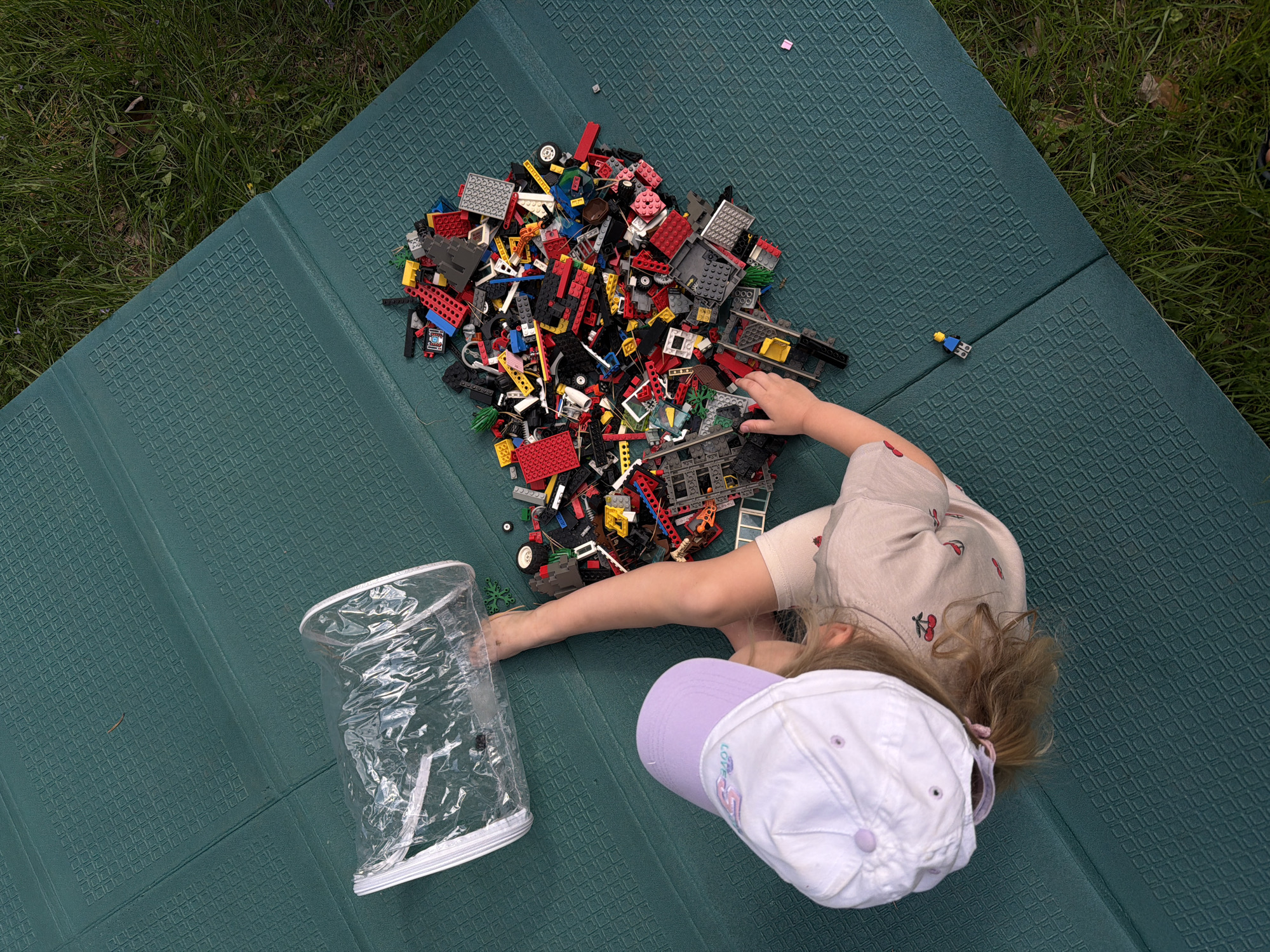A young child, wearing a white baseball cap with a lavender brim and a tan shirt with cherry designs, is sitting on a green, rectangular mat. To the child's left is a clear, zippered bag. In front of the child is a pile of Lego bricks. The setting appears to be outdoors, with grass visible in the upper left corner. The location is Udelnaya, Russia.