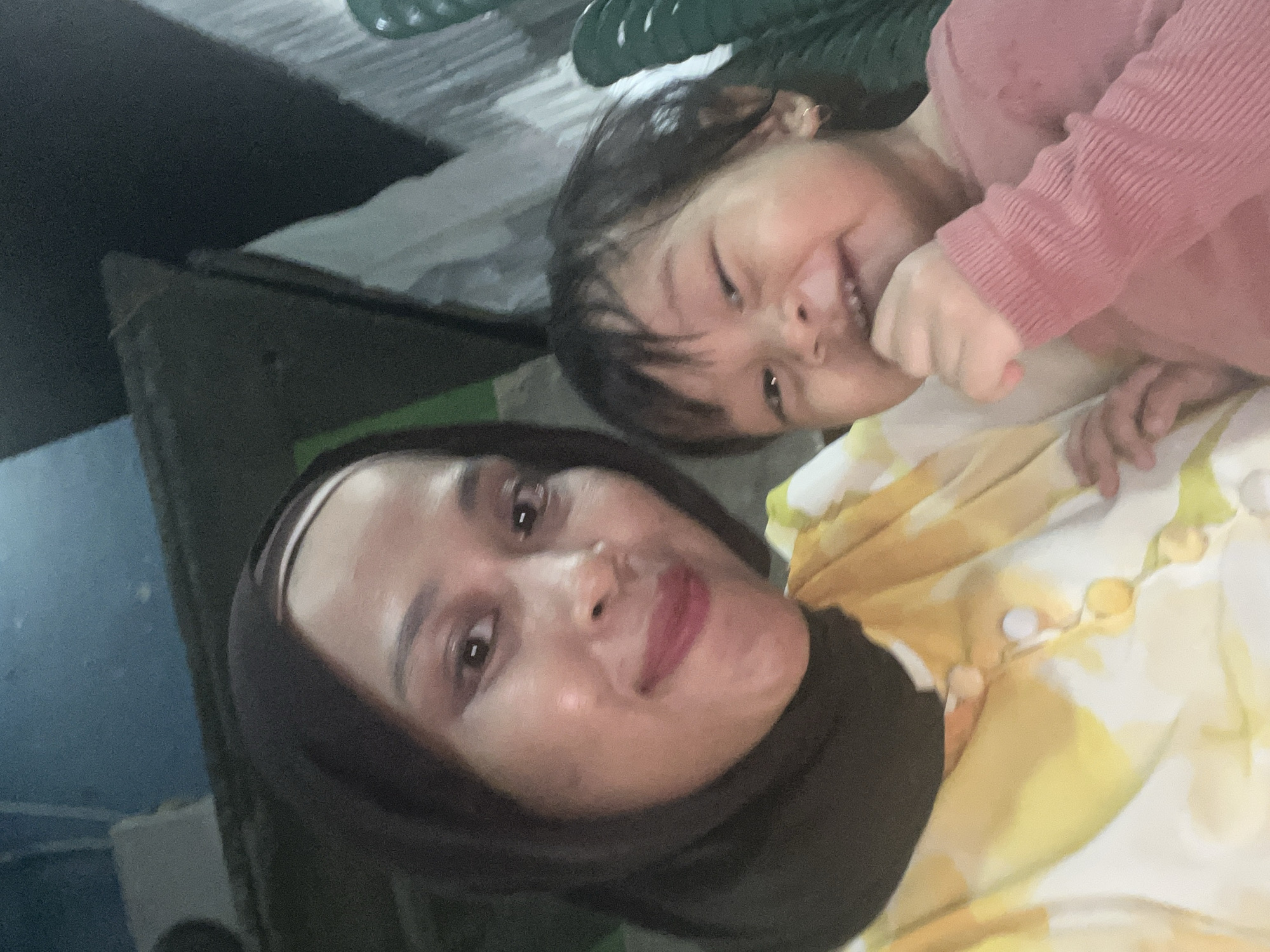 In Wedarijaksa, Indonesia, a medium shot captures a woman and a young child posing for a selfie. The woman, wearing a dark hijab and a yellow and white patterned shirt with buttons, is on the left, looking at the camera with a slight smile. Her makeup includes lipstick. To her right, a child in a pink long-sleeved shirt is smiling widely, with their hand near their mouth. The background is dimly lit and appears to be outdoors or in an open area, with hints of structures and possibly furniture. The overall impression is a casual, candid moment shared between the two individuals.