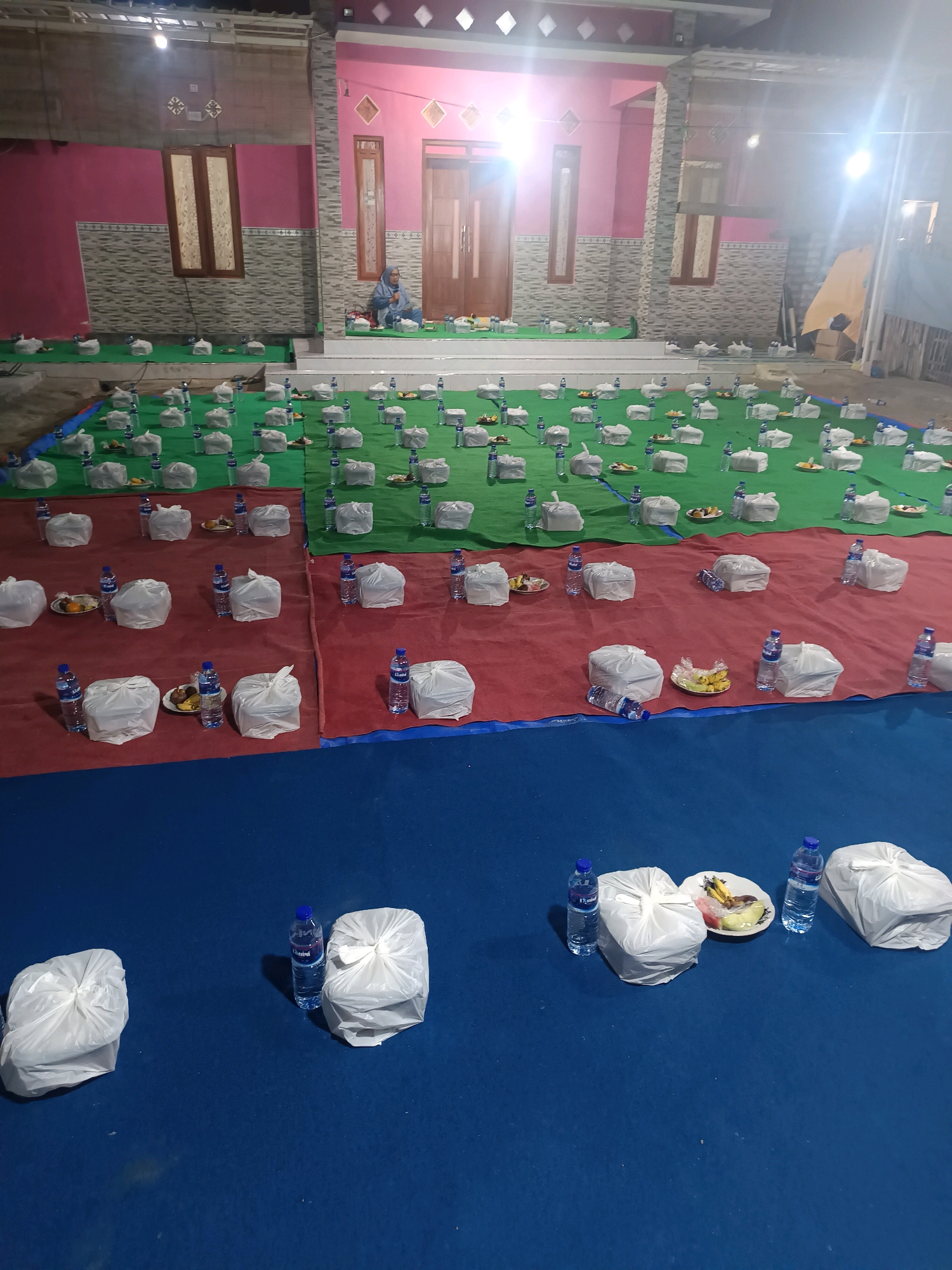 In Gunung Sekar, Indonesia, a gathering is taking place outdoors, likely a communal meal or event. The scene is set in front of a pink and grey building with a tiled facade. A woman wearing a hijab sits on a raised platform to the left of the building's entrance, with food and water bottles laid out before her.
The ground is covered with large blankets and mats in green, red, and blue. Spread across the green and red areas are numerous individually packaged meals, each consisting of white plastic bags containing food and a plastic bottle of water. Small plates of fruit, likely bananas and other items, are also visible on some of the mats. The blue mat in the foreground also has a few similar meal packages and fruit plates. The event appears to be held at night, illuminated by artificial lighting, creating bright, slightly washed-out areas. The overall impression is one of organized preparation for a shared meal.