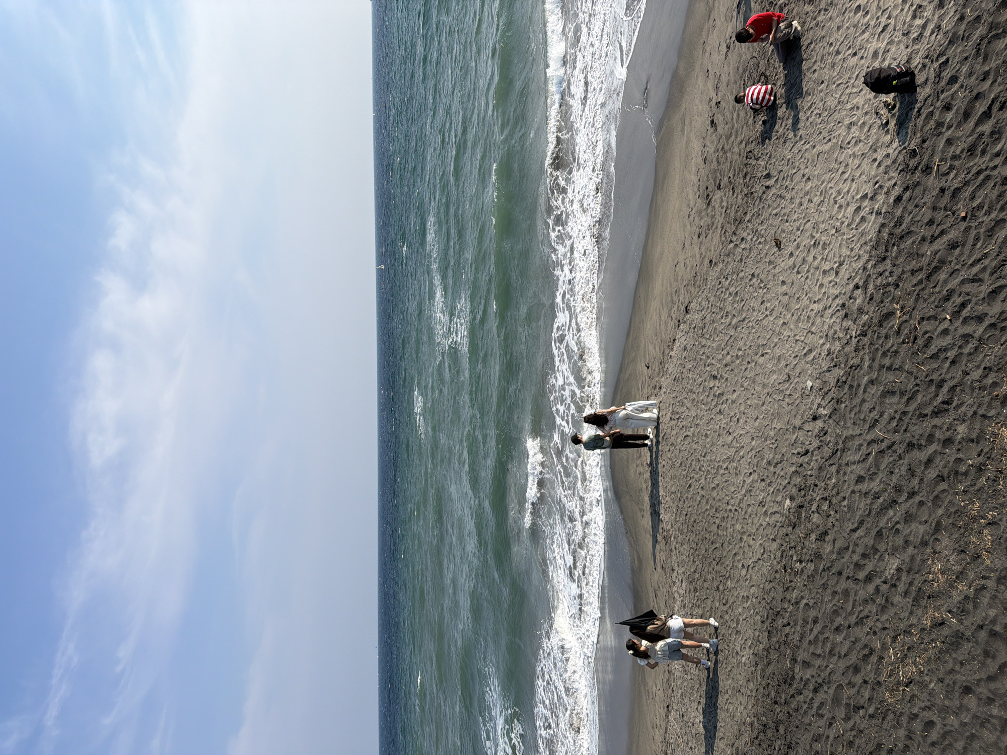 The image depicts a beach in Kamakura, Japan, with a dark sand shoreline meeting the choppy, teal-colored ocean under a bright blue sky with scattered white clouds. In the middle of the frame, near the water's edge, a couple in formal attire, presumably for a wedding photoshoot, stands facing the ocean. To their left, three young people are gathered, two of whom are looking at the couple, and one is holding a black umbrella. Further to the right on the sand, two children are playing. One child, wearing a red and white striped shirt, is seated, while another child, in a red shirt, is also seated, looking at a tablet device. A black backpack rests on the sand in the foreground. The overall scene captures a typical day at the beach with various individuals enjoying the seaside.