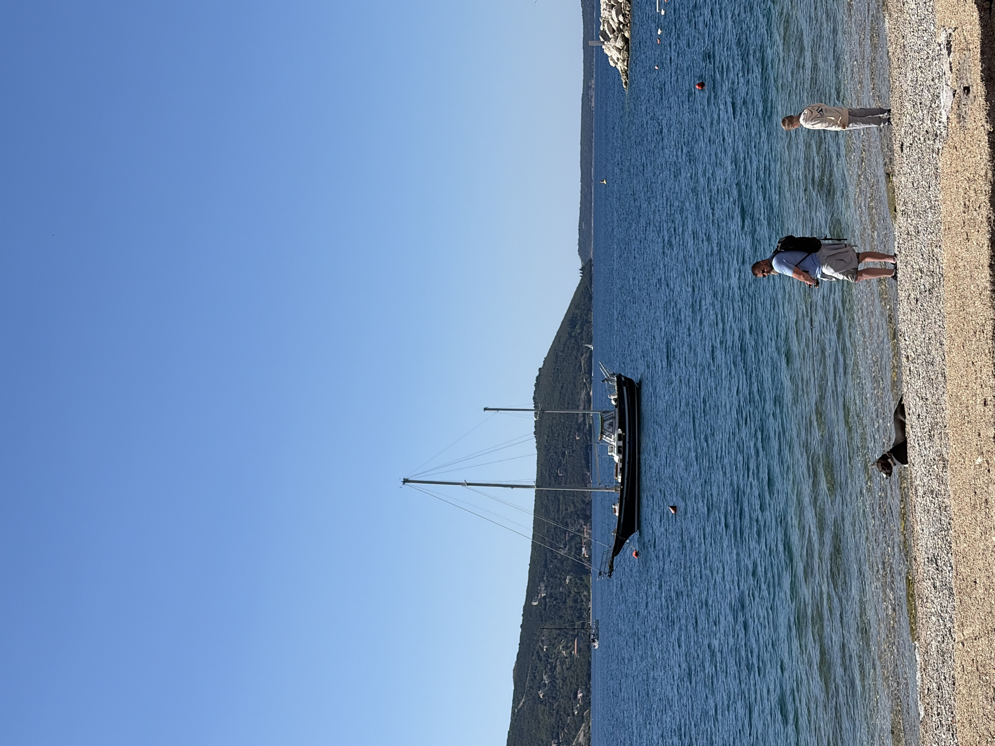 The image depicts a seaside scene in Meljine, Montenegro, under a clear blue sky. A pebble beach stretches along the right side of the frame, meeting the turquoise water of the Adriatic Sea. Two people are walking along the beach, their figures appearing relatively small in the landscape. Further out in the water, a large, dark-hulled sailing yacht is moored. The coast rises behind the yacht, covered with dense green vegetation. In the distance, the horizon is marked by a rocky breakwater. A dog stands at the edge of the water.