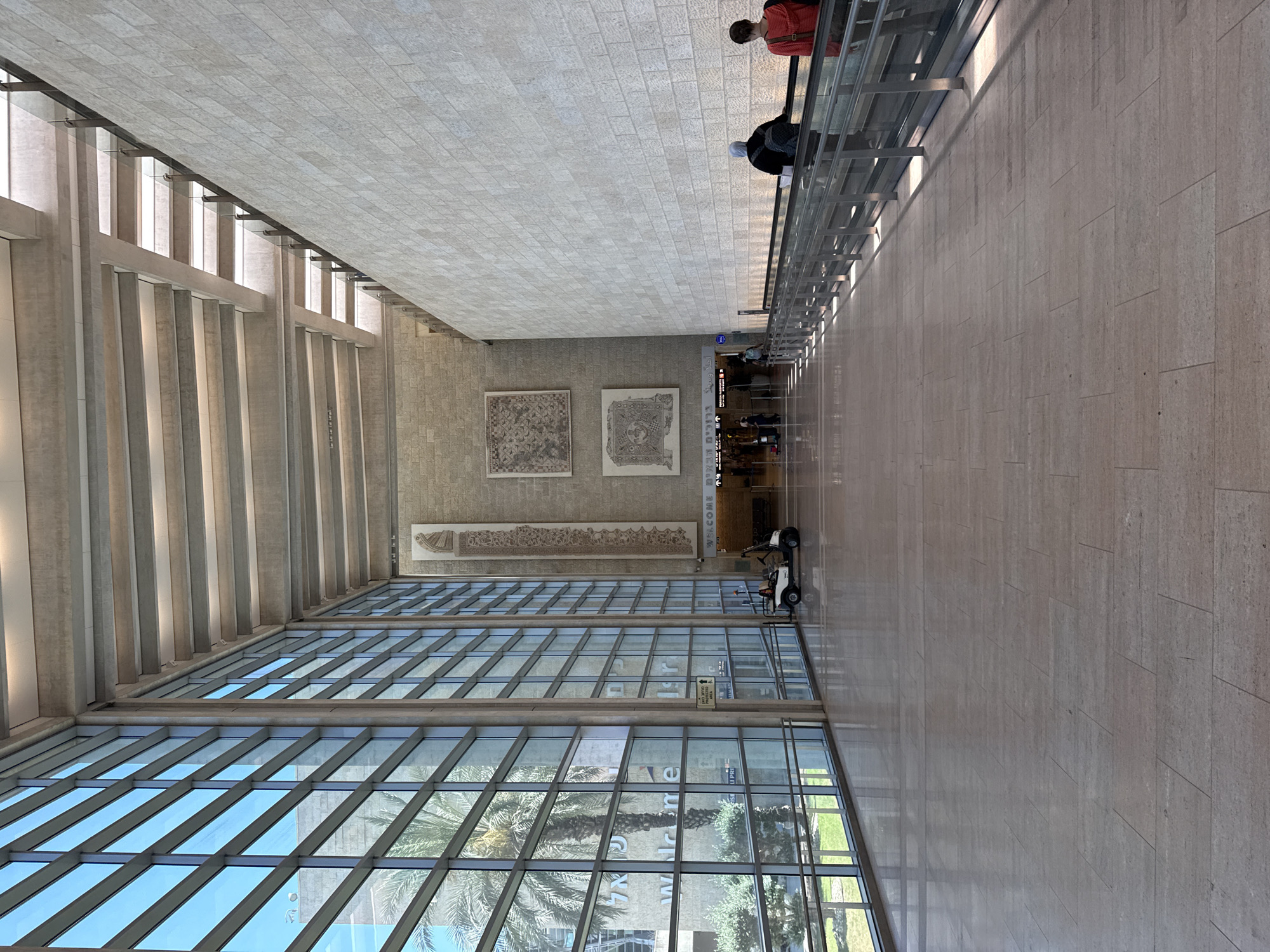 The image depicts the interior of a modern airport terminal in Tzafria, Israel. The scene is characterized by a vast, open space with high ceilings supported by concrete beams. The left side of the terminal features a floor-to-ceiling glass facade, offering a view of palm trees and greenery outside. The right side is dominated by a large, textured stone wall.
In the mid-ground, a moving walkway is visible, with two individuals, one wearing a red top and the other in a black top with a headscarf, standing on it. Further down the walkway, more people are seen, along with a small white utility vehicle. The floor is made of polished stone tiles.
On the stone wall, three large art installations are displayed. The two lower ones are square mosaics, while the uppermost one is a long, vertical rectangular tapestry. Above the entrance to a passage, a sign reads "WELCOME" in English, accompanied by Hebrew text.
The lighting in the terminal is a combination of natural light streaming through the glass windows and artificial light from the ceiling. The overall atmosphere is one of spaciousness and modern design.