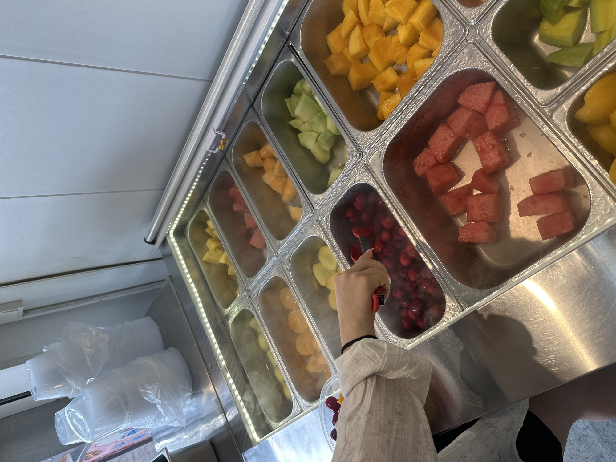 The image depicts a close-up view of a self-serve fruit bar, likely located in a food establishment within the Chongqing High-tech Zone, China. A person's hand, wearing a white, loosely woven shirt, is visible on the left side of the frame. The hand is holding red tongs, actively selecting bright red berries from a metal container filled with the fruit. The fruit bar features several stainless steel rectangular containers, each filled with a different type of chopped fruit. These include what appear to be melon chunks (light green), mango cubes (yellow), watermelon pieces (pink), and possibly orange segments. Some containers exhibit condensation, suggesting they are chilled. In the background, stacks of clear plastic cups are visible, wrapped in plastic film. The overall environment suggests a casual eatery or market stall specializing in fresh fruit.