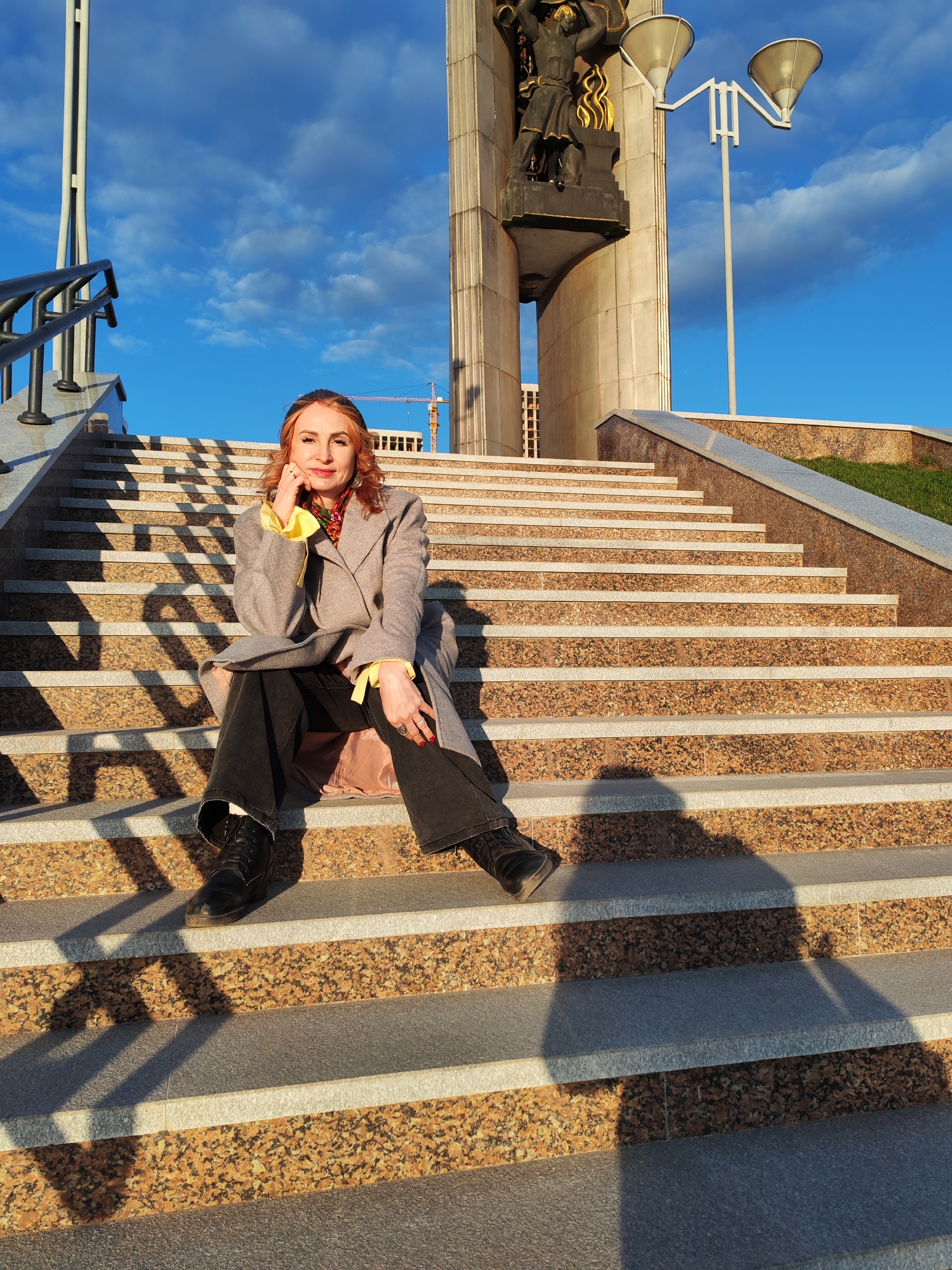 A woman sits on a wide stone staircase, posing for a photograph in Mikhailovskii, Russia. She is wearing a grey coat over a yellow blouse, black pants, and black boots. The background features a large monument with a statue of a figure holding a hammer and sickle aloft, and a clear blue sky with scattered clouds. Shadows from the setting sun are cast across the steps, adding depth and texture to the image. A crane can be seen in the distance, beyond the monument.