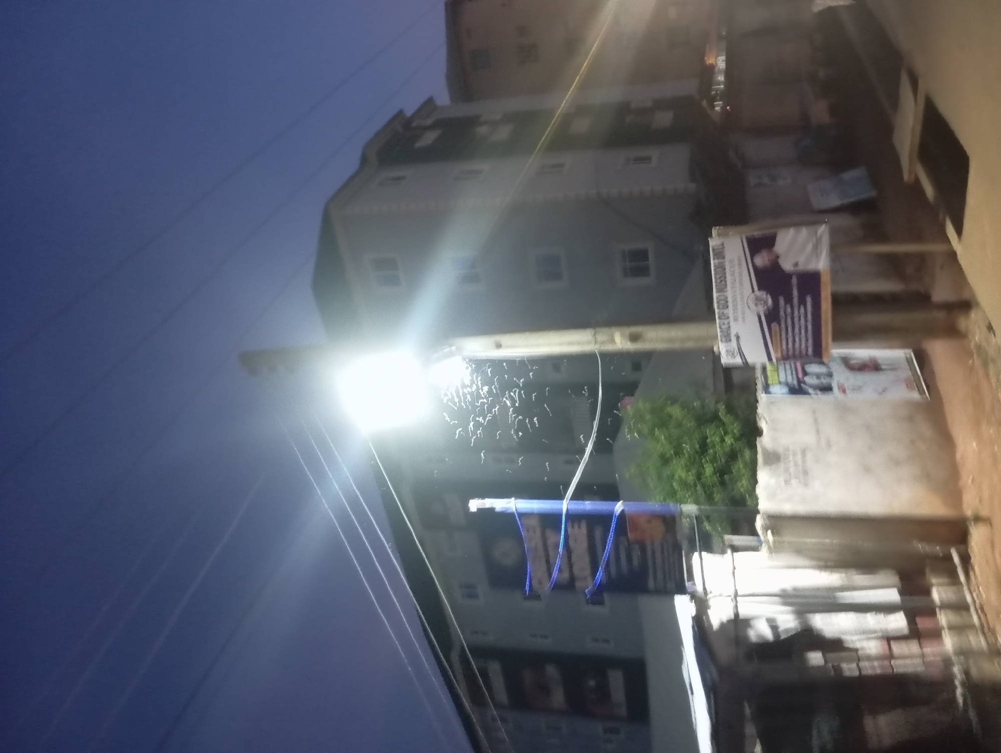 The image shows a street scene in Nkpor, Nigeria at dusk. A tall, modern building with multiple stories and windows is visible in the background. In the foreground, a bright street lamp illuminates a swarm of insects. Various signs and posters are attached to a post near the light. A blurry concrete structure is also in the foreground. Power lines are visible against the evening sky.