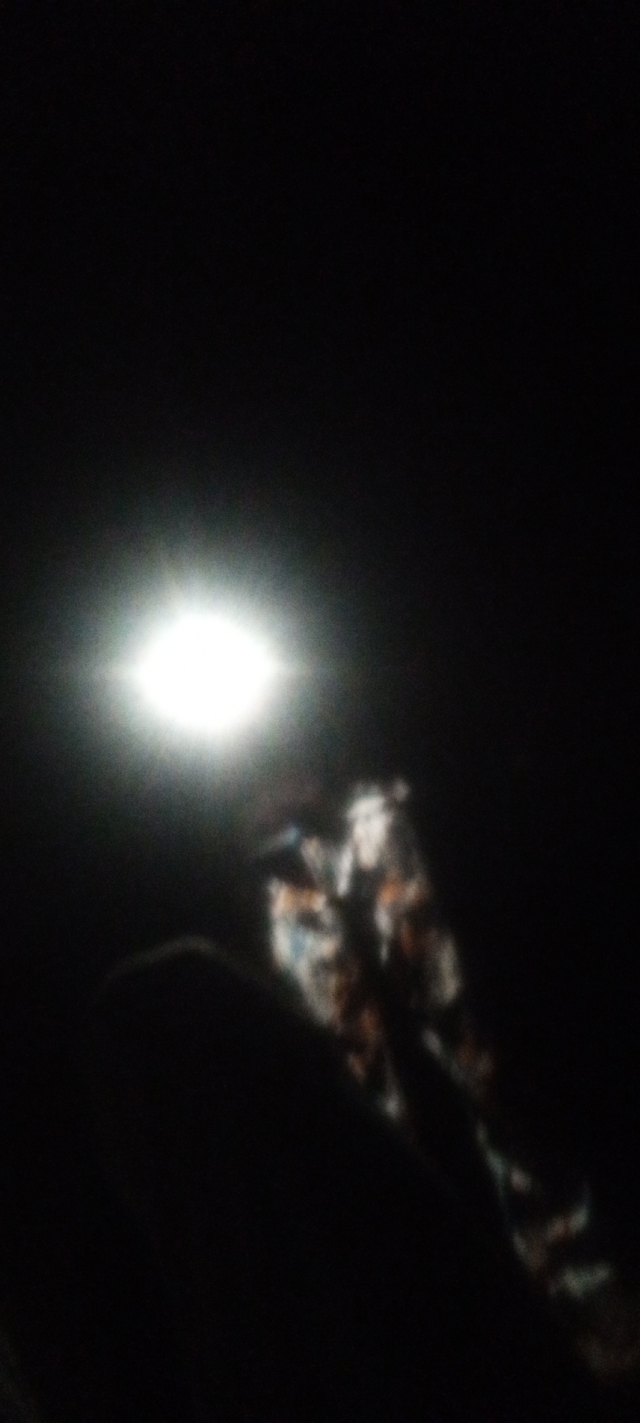 The image shows a dark scene with a bright light source in the upper left quadrant. Below the light is a blurry figure, possibly a person wearing patterned clothing, with dark silhouettes of what might be hands or arms in the foreground. The location is Unknown, Unknown, but there are insufficient details to establish anything else about the city. The scene appears to be indoors, given the limited visibility.