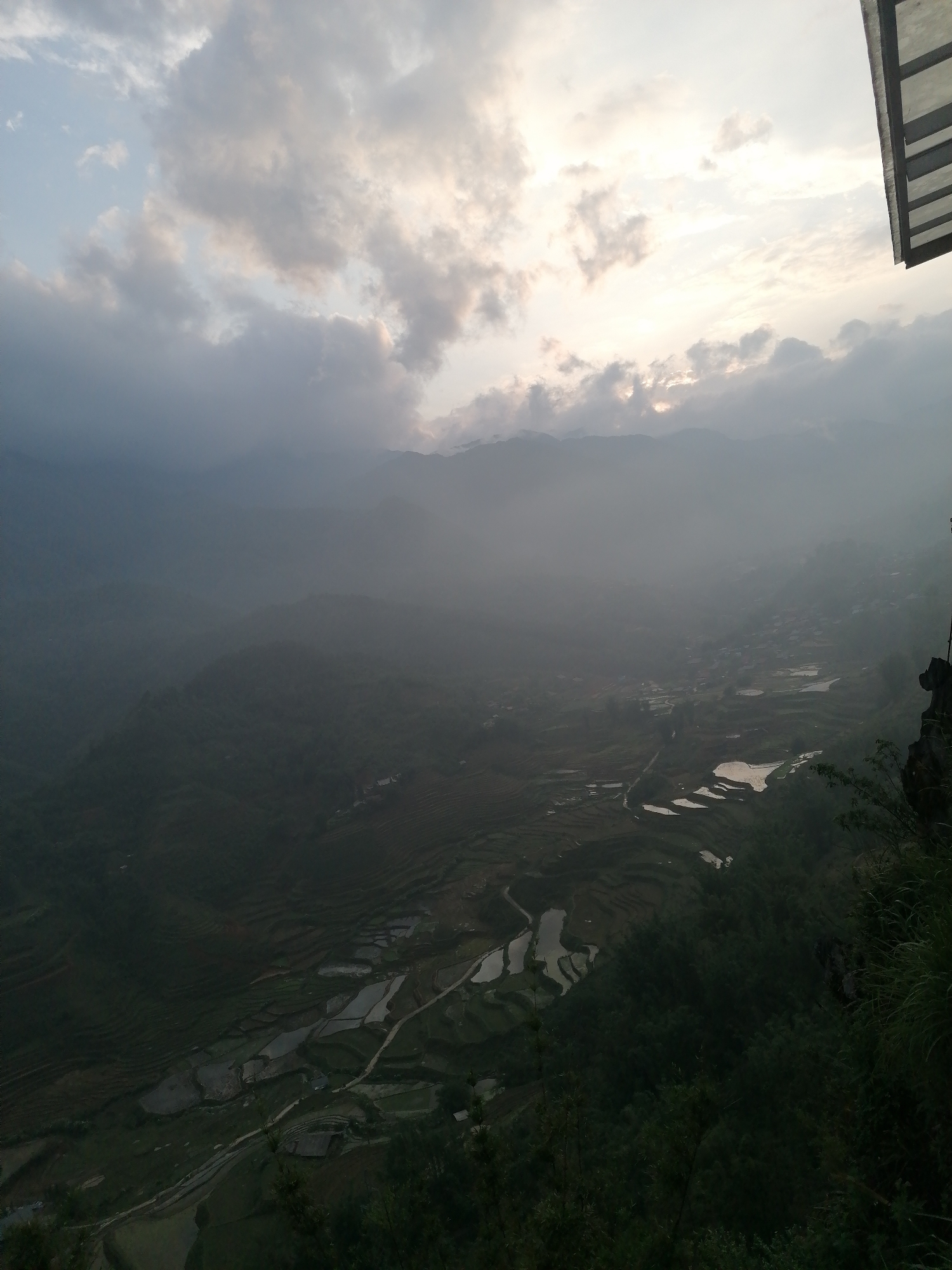 The image depicts a landscape in Sa Pa Ward, Vietnam. The scene is dominated by terraced rice paddies, partially filled with water, and nestled within a mountainous valley. A dense fog or mist blankets the lower portions of the valley, obscuring details. The sky is overcast with clouds that are tinted by the setting or rising sun. Part of a structure is visible in the top right corner, including an overhang. The perspective is from an elevated vantage point.