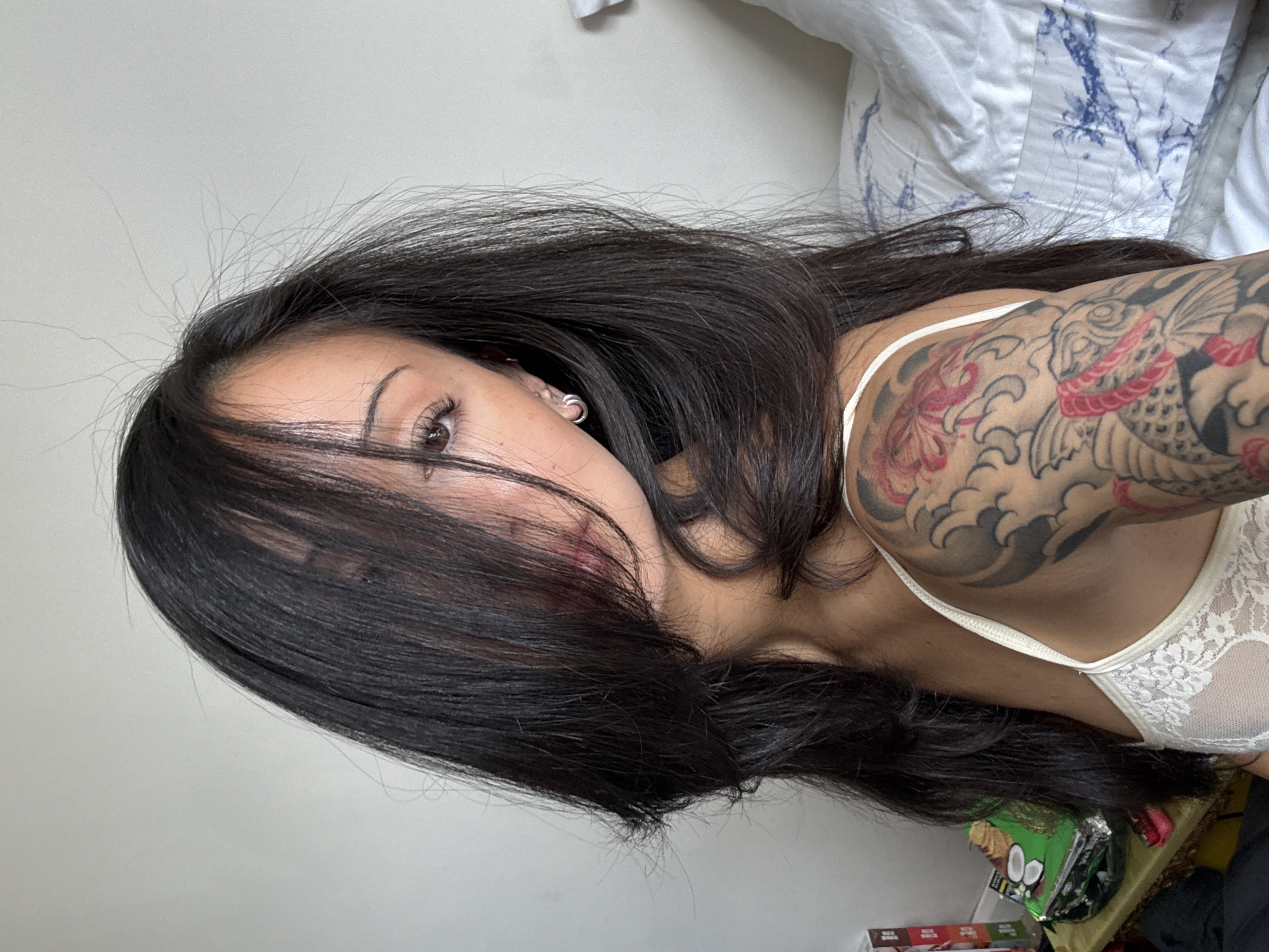 The image is a close-up, waist-up shot of a woman with long, dark hair that partially obscures her face. She is wearing a white lace camisole, and a tattoo is visible on her upper left arm. She appears to be indoors, with a white wall and a bed with blue and white patterned bedding visible in the background. To the left, there is a table with some items on it, including a green package and a red-covered object. The scene does not provide any information that can be used to determine the location within the city of London, United Kingdom.