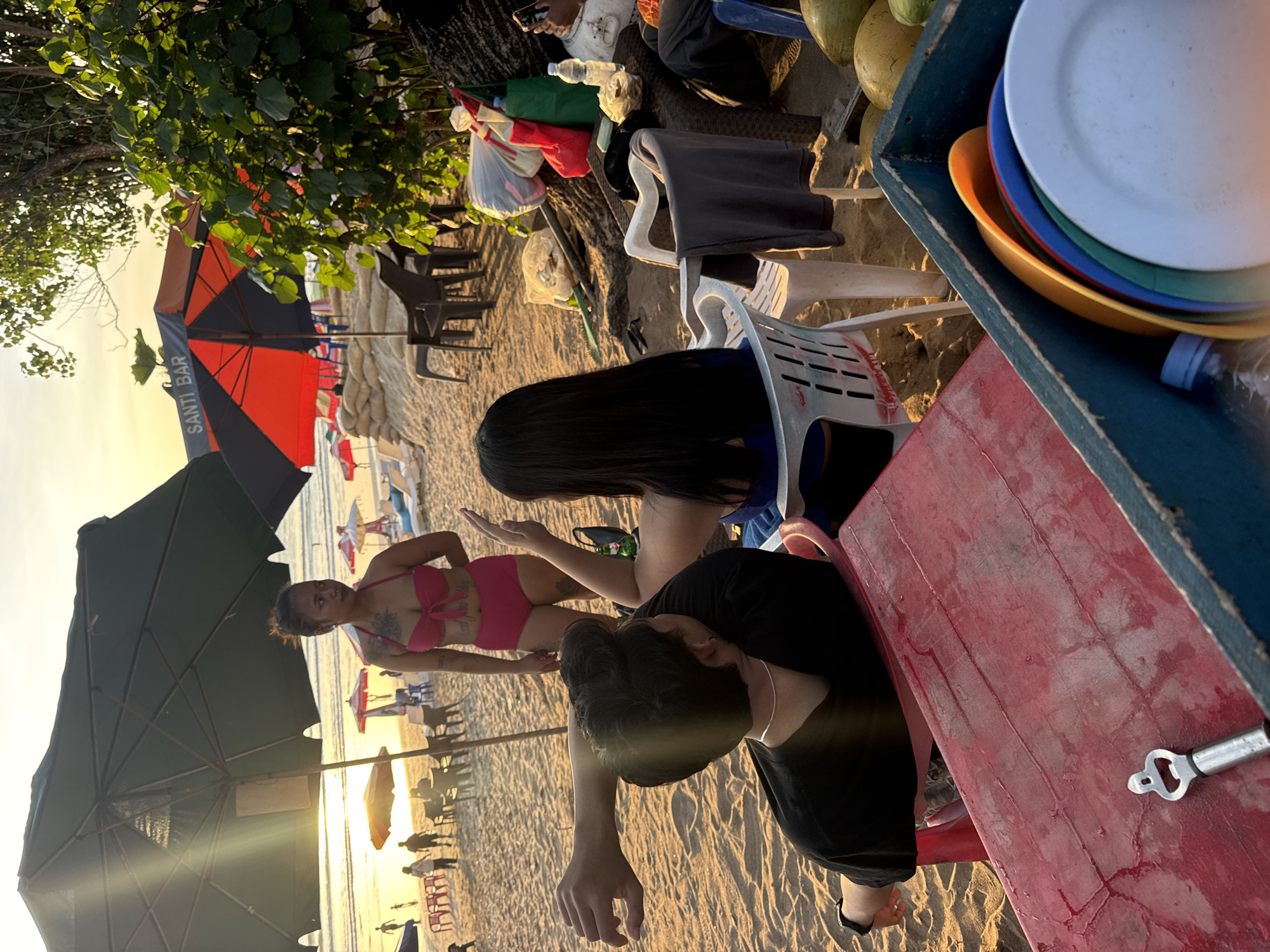 This image captures a sunset scene on Kuta Beach, Indonesia. In the foreground, a person with short, dark hair wearing a black t-shirt is seated, their left arm extended outwards. To their right, another person with long, dark hair is seated, facing away from the camera and wearing a blue top. In the mid-ground, a woman in a pink bikini stands, looking to her left. Behind her, beach umbrellas and lounge chairs dot the sandy expanse, with several people visible in the distance. To the right of the frame, lush green foliage overhangs the scene. A red plastic table with a bottle opener rests in the lower left, and a blue crate filled with colorful plates is situated in the lower right. The warm, golden light of the setting sun bathes the entire scene.