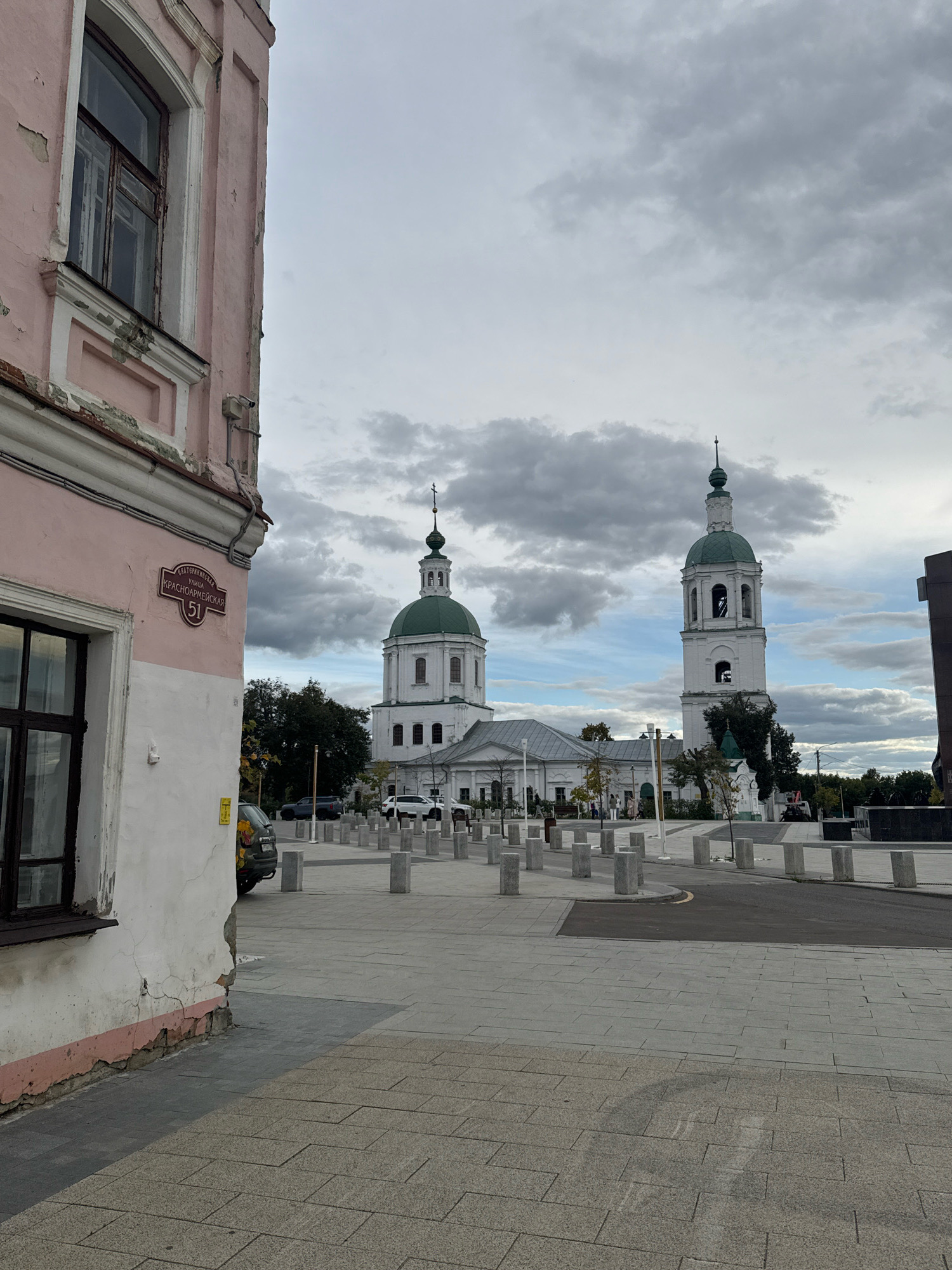 The image depicts a street scene in Zaraysk, Russia, with a focus on historic architecture. In the foreground, a weathered pink building stands on the left, exhibiting peeling paint and a prominent window. A street sign on the building reads "УЛИЦА КРАСНОАРМЕЙСКАЯ 51" (Krasnoarmeyskaya Street 51). Beyond the building, two white church towers with green domes rise against a cloudy sky. The taller tower, a belfry, features arched openings. The area between the building and the churches is paved with stone and lined with cylindrical bollards. Several cars are parked in the background, and a few individuals are visible walking near the churches. The overall environment appears to be a public square or street undergoing some form of renovation or landscaping, indicated by the uniform paving and bollards.