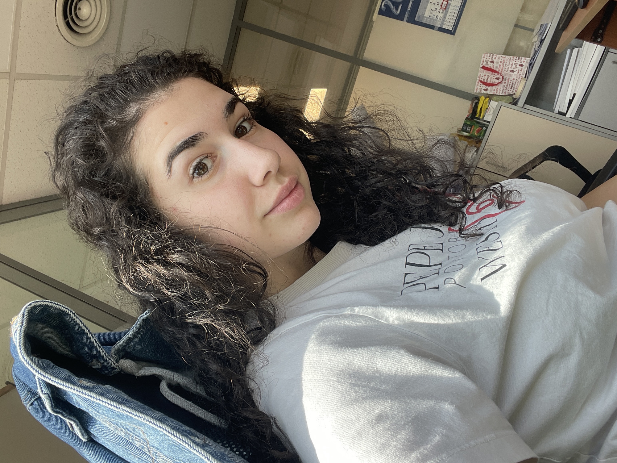 The media file presents an upward-angled portrait of a young woman in an indoor setting, likely an office or workspace, in Sofia, Bulgaria. She has dark, voluminous curly hair and brown eyes, looking towards the camera with a neutral, calm expression.
She is wearing a white t-shirt with dark lettering, and a denim jacket or similar blue fabric garment is visible in the lower left foreground. Bright natural light illuminates her face and casts shadows on her clothing, indicating it is daytime.
The background shows a ceiling with a circular vent in the upper left. To the right of the subject, a calendar with the number "2" is partially visible on a wall. Below the calendar, a small red and white gift bag with a festive pattern and a red handle is placed on a surface. Further to the right, shelves or filing cabinets containing documents are visible, along with reflections from a window or glass partition.
Visible text on her white t-shirt includes "REPE", "ROTORO", and "YVES", suggesting part of a brand name or design. The overall scene conveys a casual and relaxed moment within a functional indoor environment.