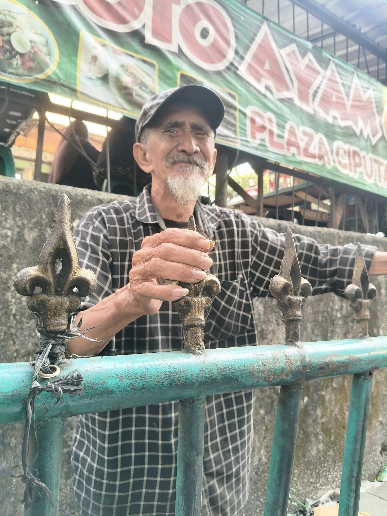 An elderly man with a goatee and a plaid shirt is standing behind a teal-colored metal fence. He is wearing a dark baseball cap. His hands are resting on the fence. A banner with red lettering is visible in the background. The scene appears to be outdoors, possibly in a street or market area. The banner text includes "AYAM" and "PLAZA CIPUTA," which suggests a location in Ciputat, South Tangerang, Indonesia, likely a food establishment selling chicken.