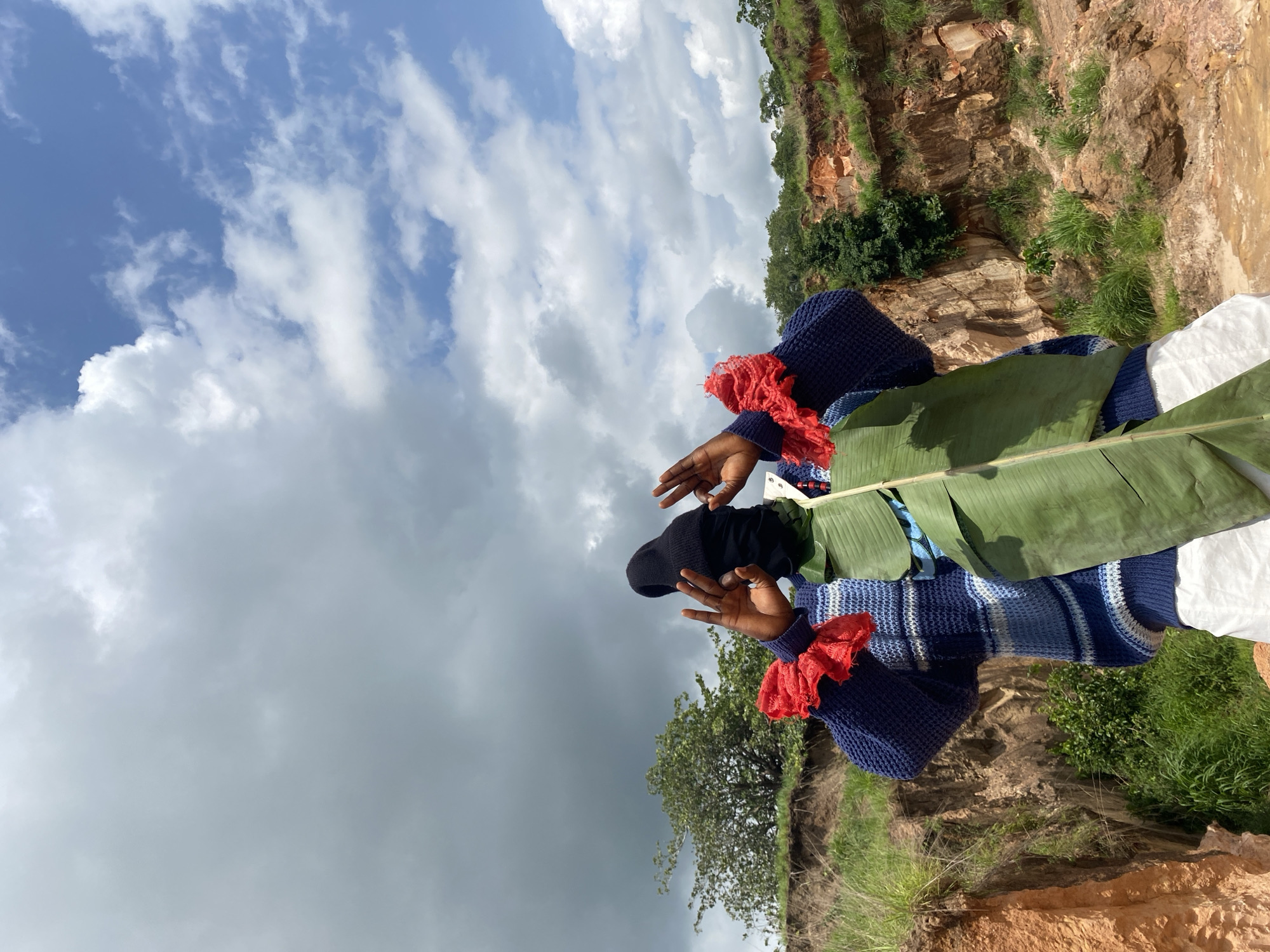 The image captures a person standing in a rugged, outdoor landscape with a dramatic sky overhead. The person is wearing a blue and white striped sweater, a black beanie, and white pants. They are holding a large green leaf in front of them and have their hands raised, fingers forming a gesture. The background features a steep, eroded embankment with exposed reddish-brown earth and green vegetation. The sky is partly cloudy, with areas of bright blue interspersed with large, dark gray clouds, suggesting changing weather. The overall scene is set in a natural, possibly remote location within the city of Ufu Esioni, Nigeria.
