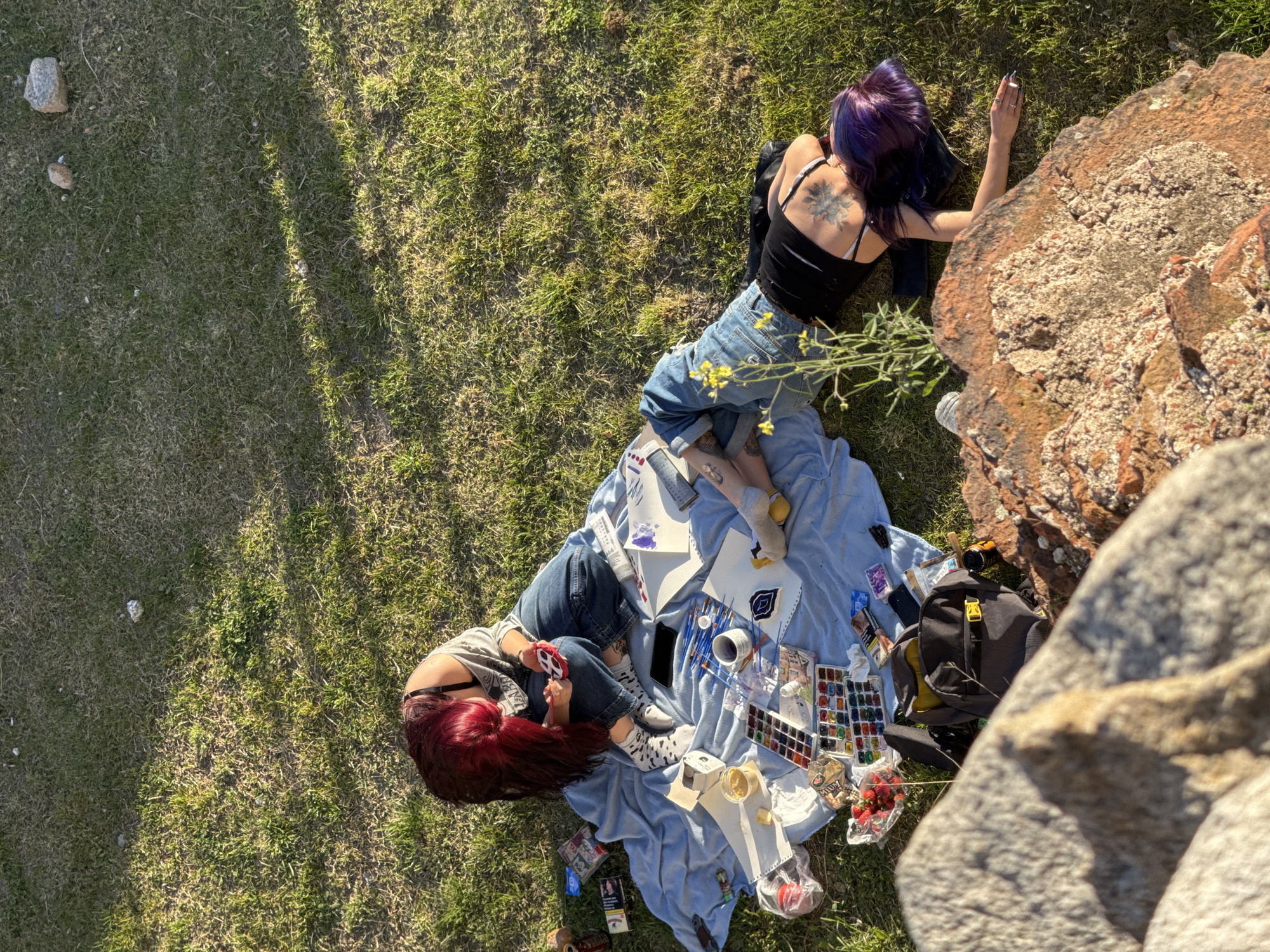Two women are laying on a blue blanket in a grassy area. One woman, with dark purple hair and a tattoo on her back, is laying partially on the blanket and partially on the grass, facing away from the camera. The other woman, with red hair, is laying on the blanket facing the camera. The blanket is covered with art supplies, including paint, brushes, paper, and strawberries. A black backpack is next to the blanket. A rock formation is visible on the right side of the image. The location is likely Nebet Tepe, Plovdiv, Bulgaria.