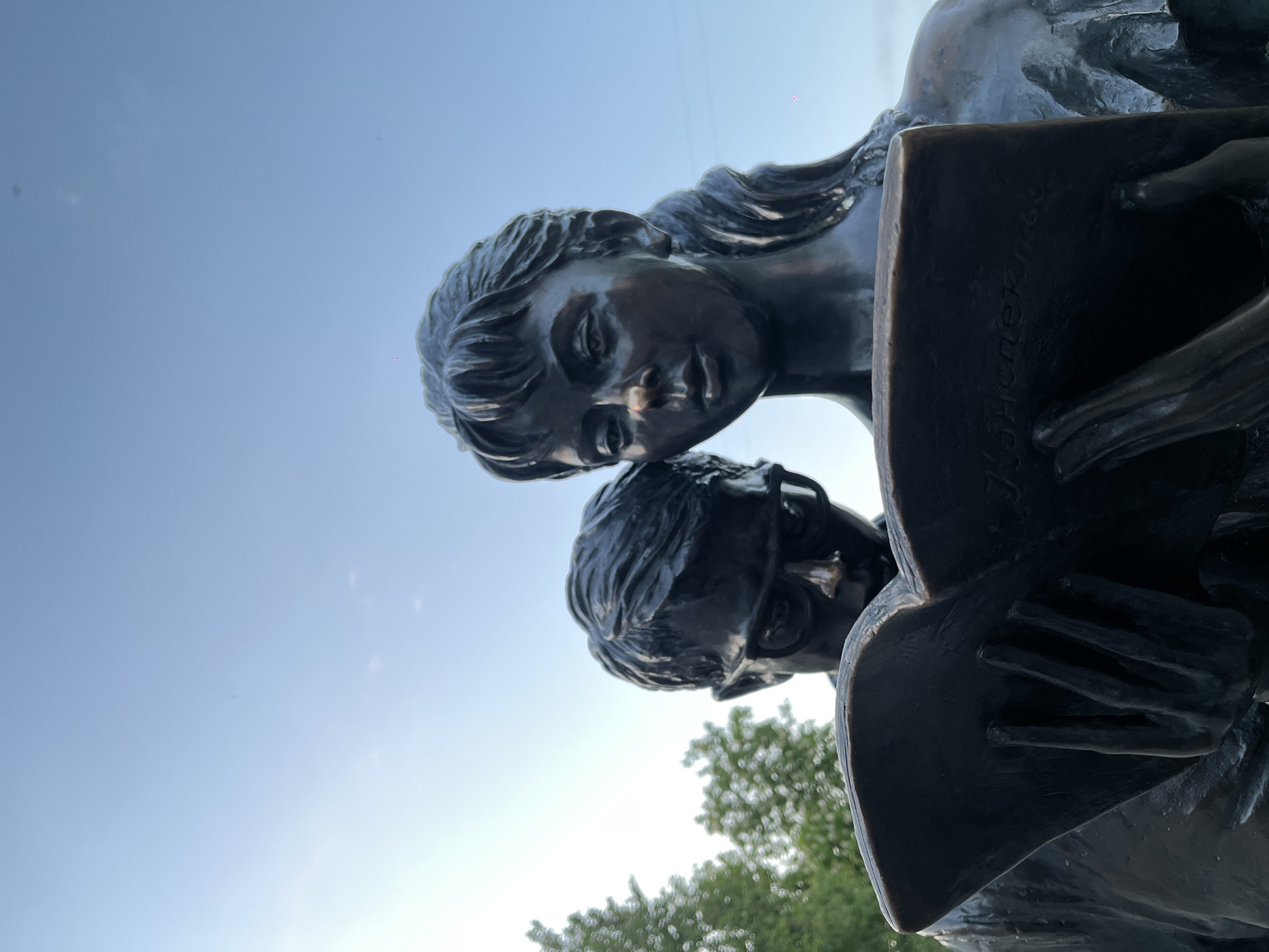 The image displays a low-angle view of a bronze sculpture featuring two young individuals, a female and a male, engaged in reading. The statues are set against a clear, bright blue sky, indicating daytime.
The female figure, positioned behind and slightly above the male, has detailed, flowing hair and a focused, soft expression as she gazes downwards. The male figure, wearing glasses, is shown holding an open book or tablet in his hands. Both statues appear intently focused on the contents of the item he holds, suggesting a shared act of reading or study. His hands are sculpted around the edges of the book.
A distinctive detail is the vertical text engraved on the book. The visible Cyrillic inscription reads "БИБЛИОТЕК...", which translates to "LIBRAR..." or "LIBRARY".
In the background, blurred green foliage, consistent with trees, is visible at the lower right of the frame, confirming an outdoor setting, likely a public park or square in Krasnodar, Russia. The bright, cloudless sky indicates clear weather. The overall scene conveys a sense of intellectual curiosity and shared concentration.