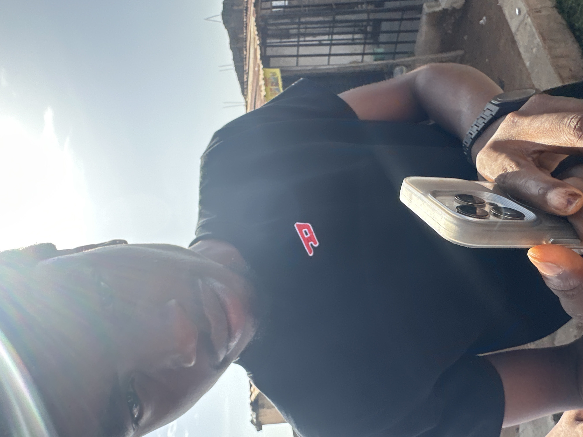 A man wearing a black t-shirt with a red "P" logo is holding a smartphone. He is also wearing a black watch. The photo appears to be taken outdoors in a city, possibly in Unknown, Unknown. There is a building structure visible in the background.