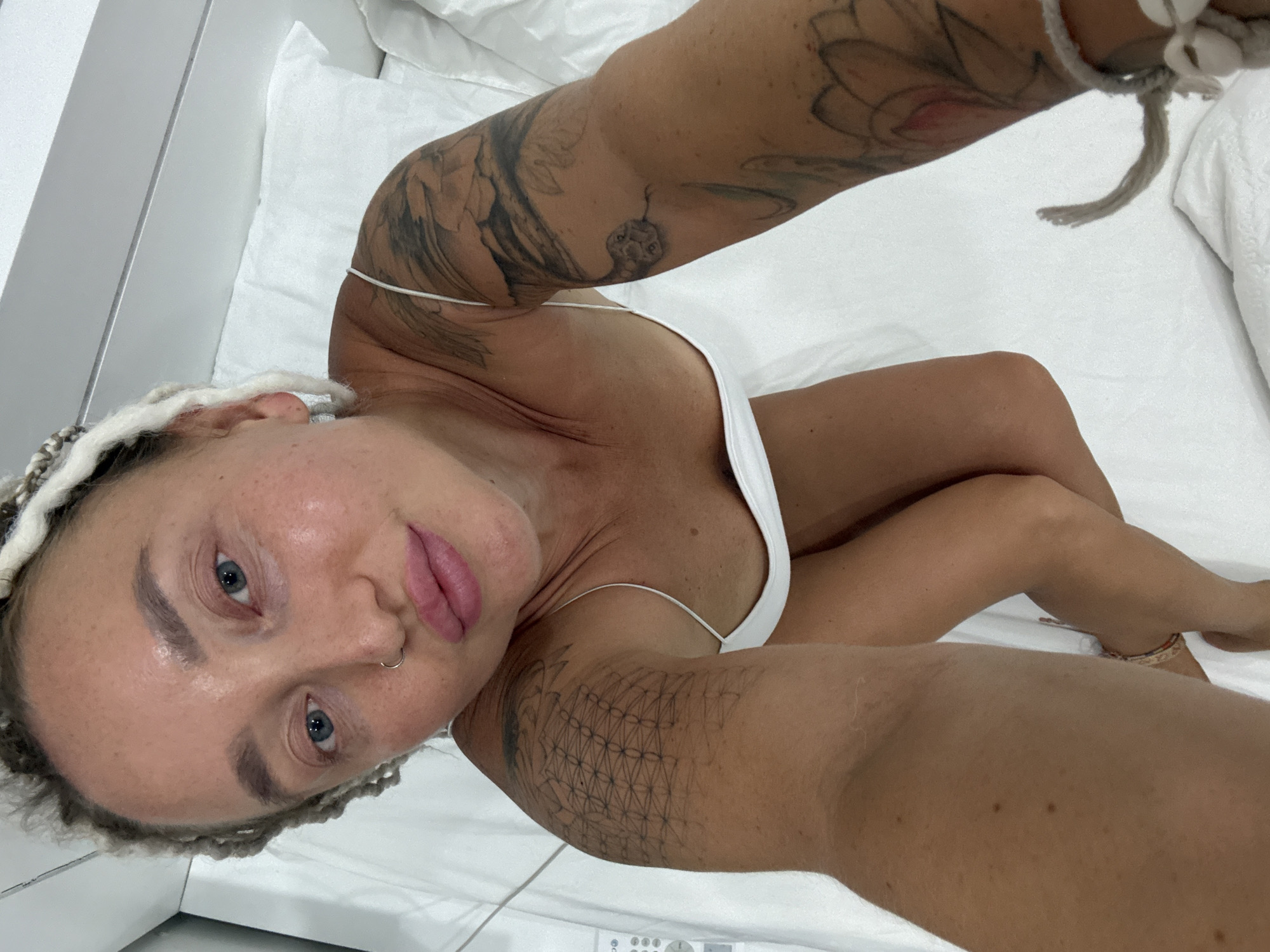 A top-down, medium shot captures a woman with blonde braided hair, wearing a white spaghetti-strap top, lying on a white surface that appears to be a bed or examination table. She has blue eyes, a nose ring, and prominent pink lips. Her arms are adorned with tattoos, one depicting intricate geometric patterns and the other featuring floral designs and a snake. Her legs are bent, and a beaded bracelet is visible on her right ankle. The environment is minimalist, with a white wall or partition visible in the background and a control panel with buttons on the left side of the frame. The lighting is bright and appears to be artificial. The woman is the sole subject of the image.