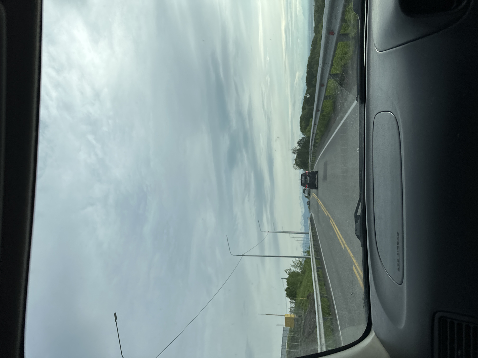 The image is a first-person perspective from inside a car, looking out through the windshield at a cloudy sky and a highway. The car is moving along a two-lane road with yellow dividing lines and a white edge line. On the left side of the road, there is a metal guardrail, a grassy embankment, and then what appears to be a construction site with a fence and a yellow structure. Several streetlights are visible along the road. Ahead, there are other cars on the road, including a dark-colored SUV. The landscape beyond the road is rolling hills covered in green vegetation, with distant mountains visible under the overcast sky. The dashboard of the car is visible in the foreground, with the text "SRS AIRBAG" imprinted on the passenger side. The scene suggests a drive through a somewhat rural or suburban area.
