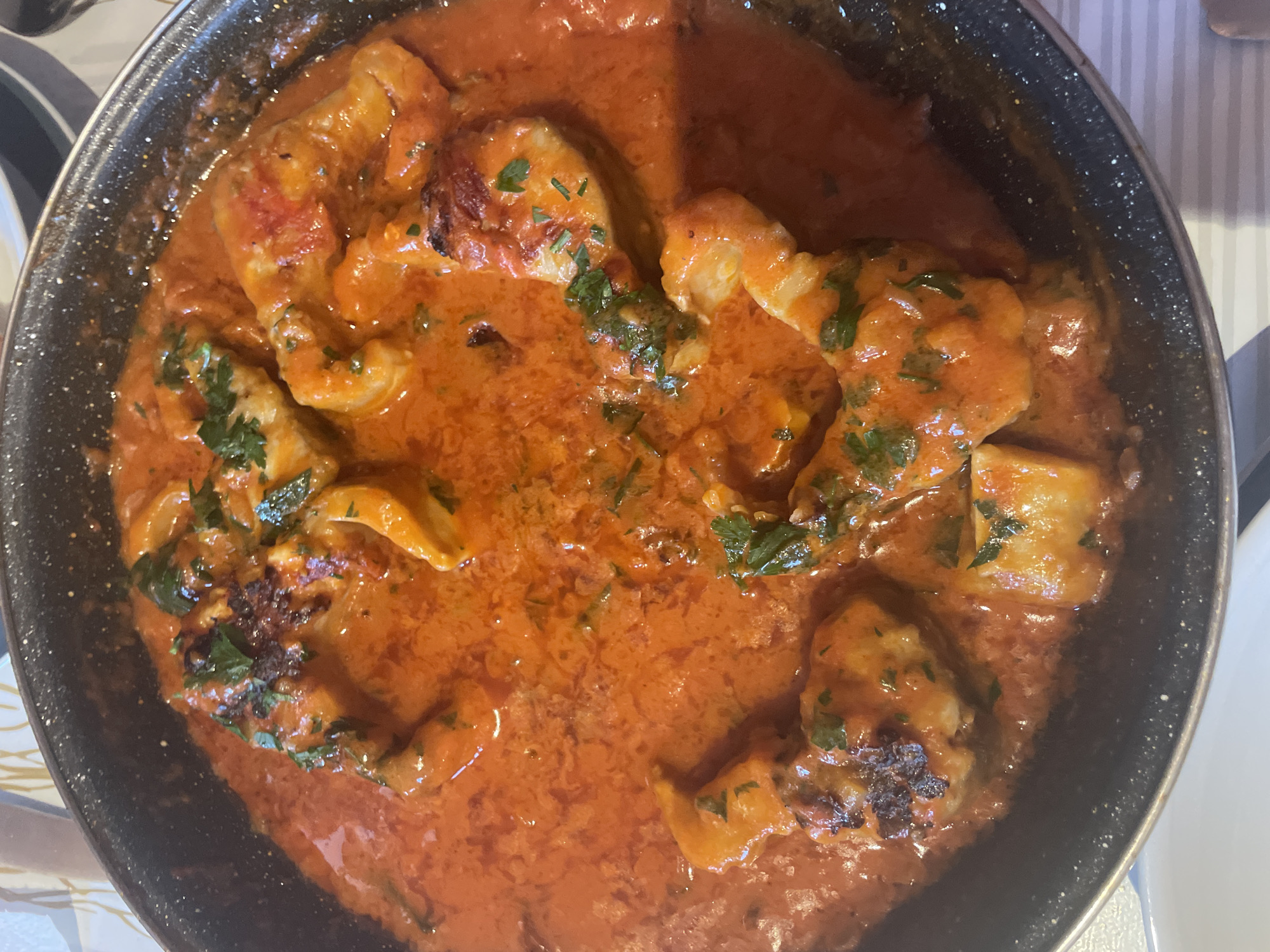 This is a close-up, top-down shot of a black, speckled pan filled with a creamy, orange sauce containing chunks of white fish and sprinkled with chopped green herbs. The fish pieces vary in size and shape, with some showing slightly seared edges. The sauce appears rich and thick, coating the fish generously. The overall impression is that of a hearty, flavorful dish, likely prepared or served recently. The background is blurred, but hints of a striped tablecloth and a white surface suggest a dining setting. The lighting appears to be indoor, with a warm cast, possibly from overhead lights. No people are visible in the frame. The context of Punta Ala, Italy, suggests this might be a local seafood preparation. There is no visible text in the image.