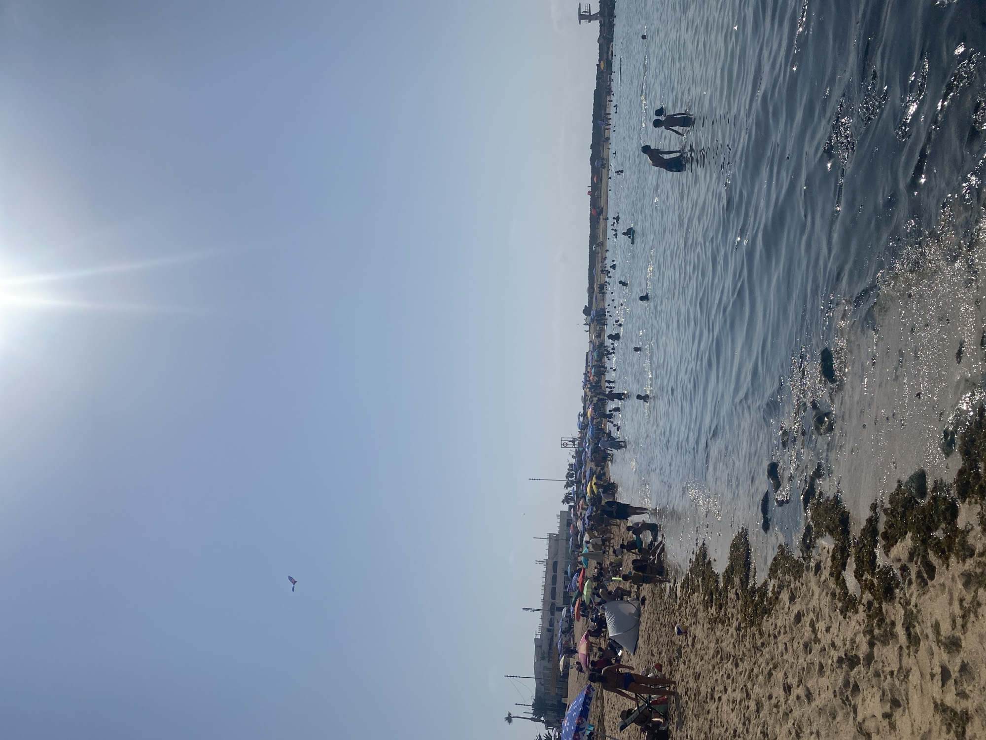 This is a full-frame, eye-level, outdoor shot of a crowded beach on a sunny day in Harhoura, Morocco. The image captures a bustling scene with a sandy beach on the left and the ocean on the right. Numerous people are scattered across the beach and in the water, enjoying the weather. On the sand, many beachgoers are seated under colorful umbrellas or relaxing on towels. Some are wading in the shallow water near the shore, while others are swimming further out. The ocean is calm with gentle waves, and the water is a deep blue. In the distance, along the coastline, there are buildings and structures, suggesting an urban or resort area. The sky is clear blue with the sun shining brightly, causing lens flare in the upper right portion of the image. A kite is visible in the sky in the upper left corner. The overall impression is one of a lively and popular beach destination.
