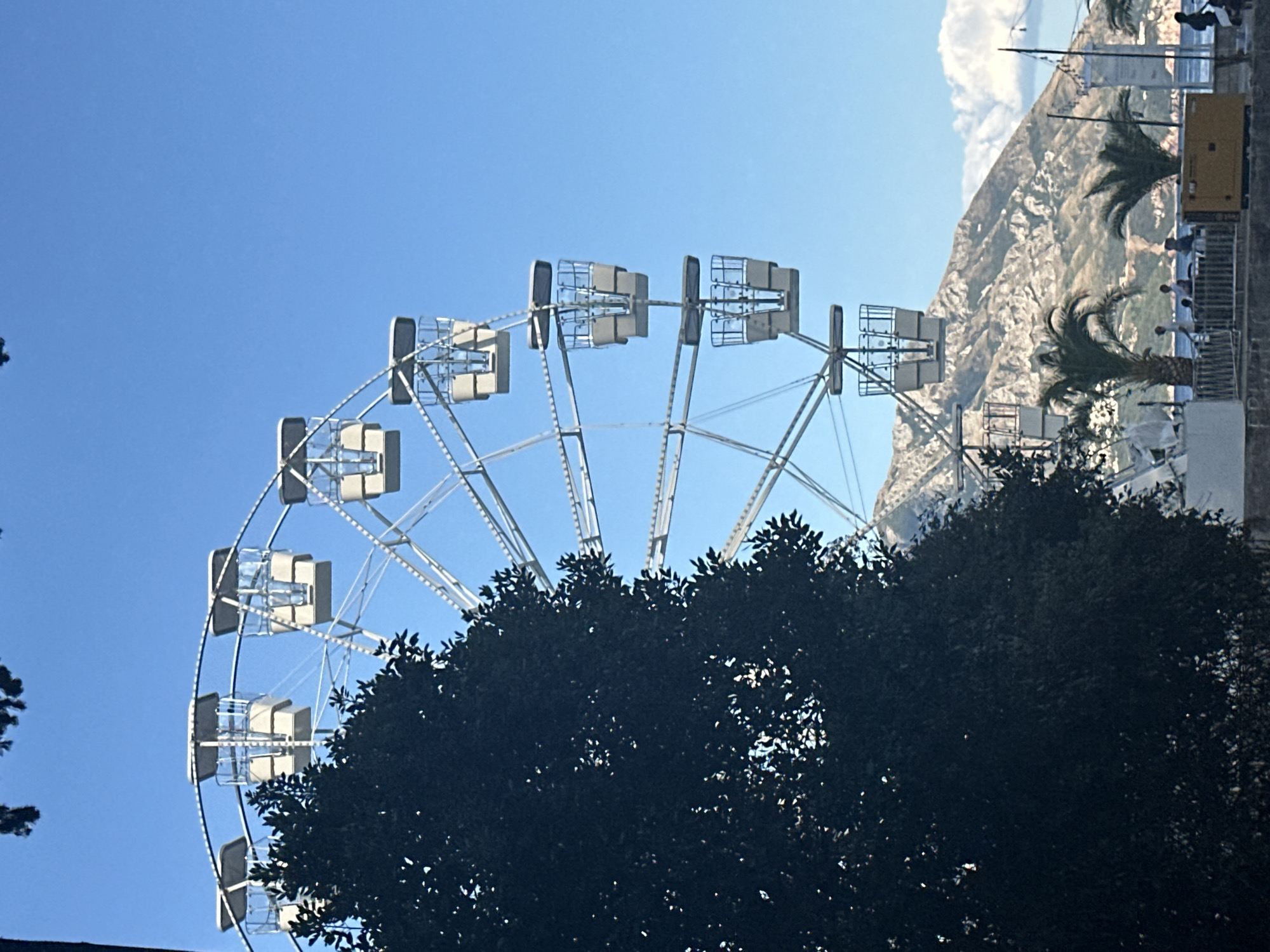 In Budva, Montenegro, a Ferris wheel stands against a clear blue sky, its white gondolas suspended from a silver frame. A large, dark green tree dominates the left foreground, its leaves obscuring part of the wheel and the background. In the mid-ground, a rugged, rocky mountain range rises behind the Ferris wheel, its slopes dotted with sparse vegetation. At the base of the mountain, several palm trees are visible, and a body of water, likely the Adriatic Sea, stretches out. A yellow generator unit and a few people are scattered along a waterfront area, some standing near a railing. The scene captures a bright, sunny day, with the Ferris wheel and the natural landscape of the coastline creating a picturesque setting.