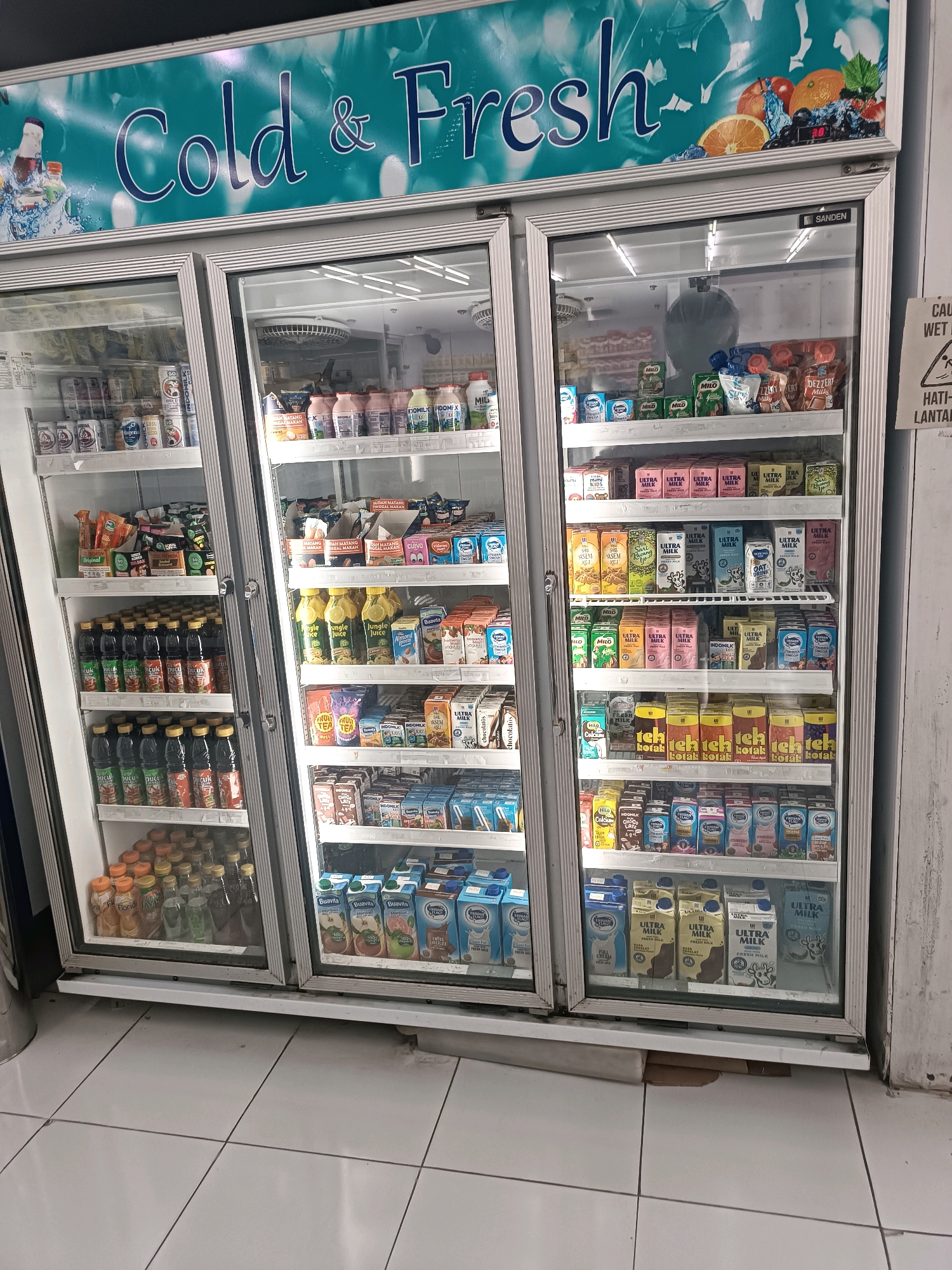 The image shows a commercial refrigerator filled with various beverages and dairy products. The refrigerator has glass doors and is divided into multiple shelves. The branding on the top of the refrigerator reads "Cold & Fresh." The products visible include milk cartons, juice bottles, and other packaged drinks. The shelves are organized with products neatly arranged. The overall scene appears to be an indoor retail environment, likely a convenience store or a supermarket, given the presence of the refrigerator displaying goods for sale. A sign on the right side of the image indicates a location in Tabanan, Indonesia, with text including "CAUTION WET FLOOR" and "HATI-HATI LANTAI LICIN." The floor in front of the refrigerator is tiled. There are no people visible in the image.