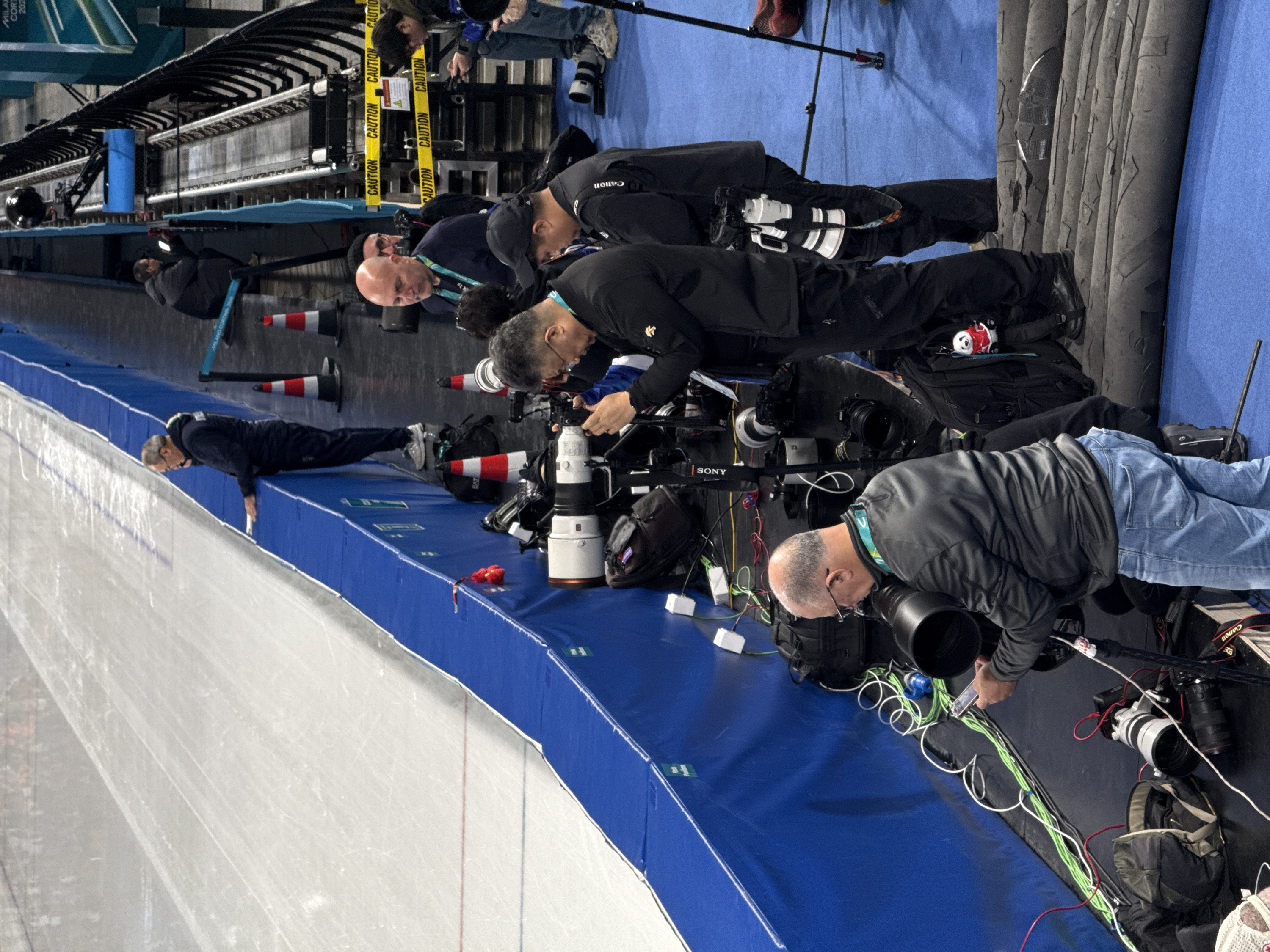 The image captures a scene from an indoor ice rink, focusing on a line of professional photographers and videographers positioned along the edge of the ice, apparently covering a sporting event. The image is oriented vertically, showing the ice rink on the left and the media personnel and background structures on the right.
In the foreground, a large expanse of white ice is visible, marked with subtle skate tracks and faint red and blue lines, indicating it's an active competition area. Separating the ice from the media area is a blue protective padding, which runs horizontally across the frame.
Along this blue padding, several men, mostly dressed in dark athletic wear like jackets and pants, are actively engaged in photography and videography. Most are focused intently towards the ice, operating their equipment.
- On the far left, one man in a dark tracksuit is lying prone on the blue padding, looking over the edge, suggesting he is capturing a low-angle shot.
- To his right, a group of photographers are either kneeling or standing, each with sophisticated camera setups. These setups include large camera bodies with prominent telephoto lenses, many of which are white or black. Brands like "Canon" and "Sony" are discernible on some of the equipment.
- The photographers have a variety of accessories around them, including black camera bags, tripods, monopods, and an array of cables (green, white, red, black) and power bricks, indicating a complex and powered media setup.
- One photographer, who is bald and wearing glasses and a dark jacket, is seen kneeling and adjusting his camera. Another man, with grey hair and glasses, is also kneeling, operating a camera with a large black lens.
- A few red and white striped traffic cones are placed in the media area.
- Notable details among the equipment include a small red object, possibly a ribbon or flower, on the blue padding, and a small panda plush toy wearing a red outfit, sitting on top of a black camera bag.
In the background, behind the photographers, a dark, ribbed or corrugated structure is visible, which could be part of the stadium seating or a temporary media platform. Yellow "CAUTION" tape with repeated text is strung across this area. Further back, more individuals and tripods can be seen, blending into the darker background. Light blue banners or stands featuring the text "MILANO CORTINA 2026" are visible in various parts of the background, strongly suggesting the event is related to the 2026 Winter Olympics.
The lighting is artificial and bright, typical of an indoor sports arena. The overall mood is one of focused activity and professional concentration, with the photographers intently capturing the action on the ice.