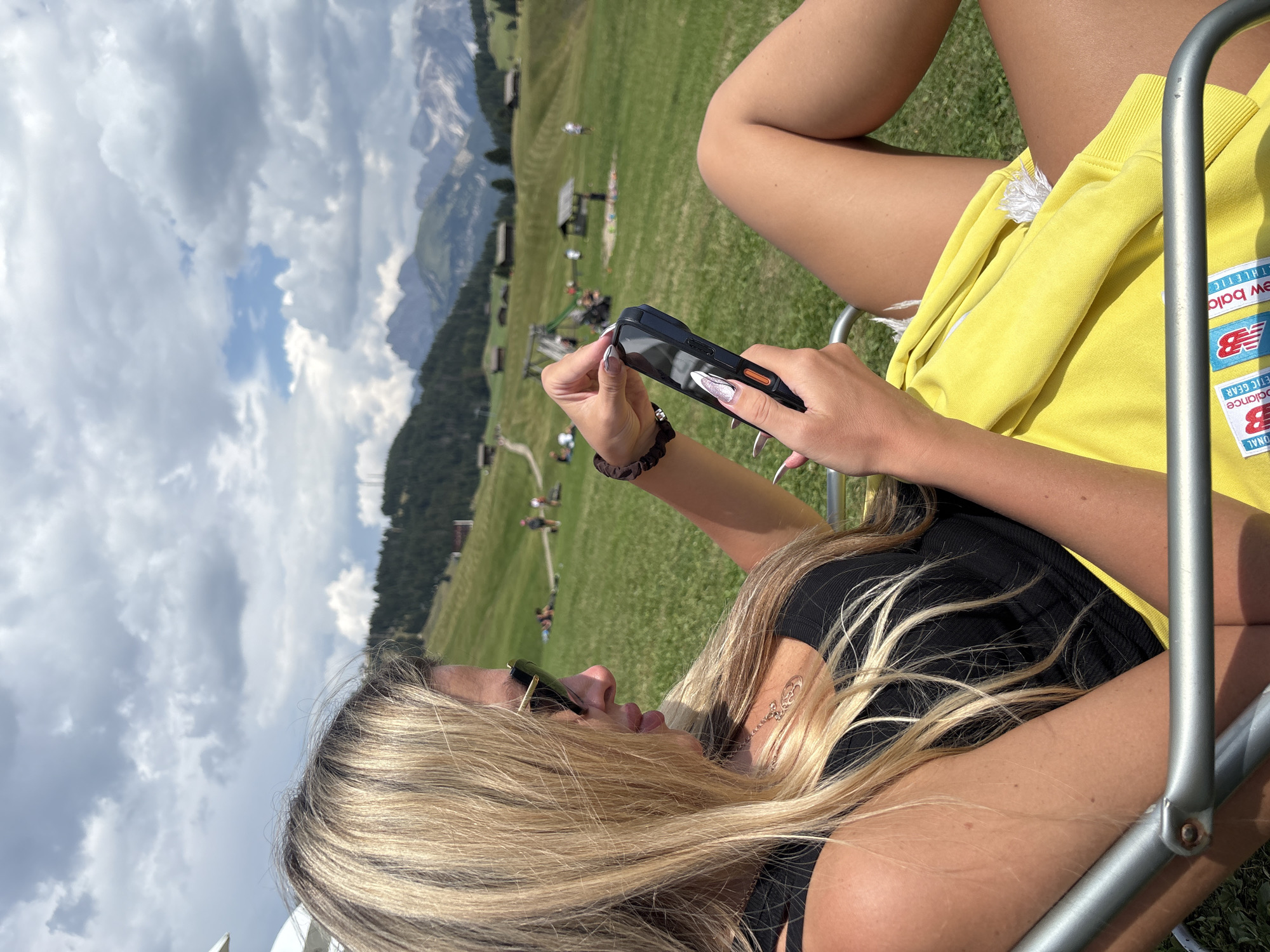 A blonde woman with long hair and sunglasses is sitting in a reclining chair outdoors, holding a smartphone in her hands and looking at the screen. She is wearing a black tank top and a yellow garment. In the background, a green grassy hillside is visible with scattered trees, small buildings, and distant mountains under a cloudy sky. The location is likely an alpine meadow near Kastelruth (Castelrotto), Italy, suggested by the characteristic architectural style of the buildings and the mountain landscape.
