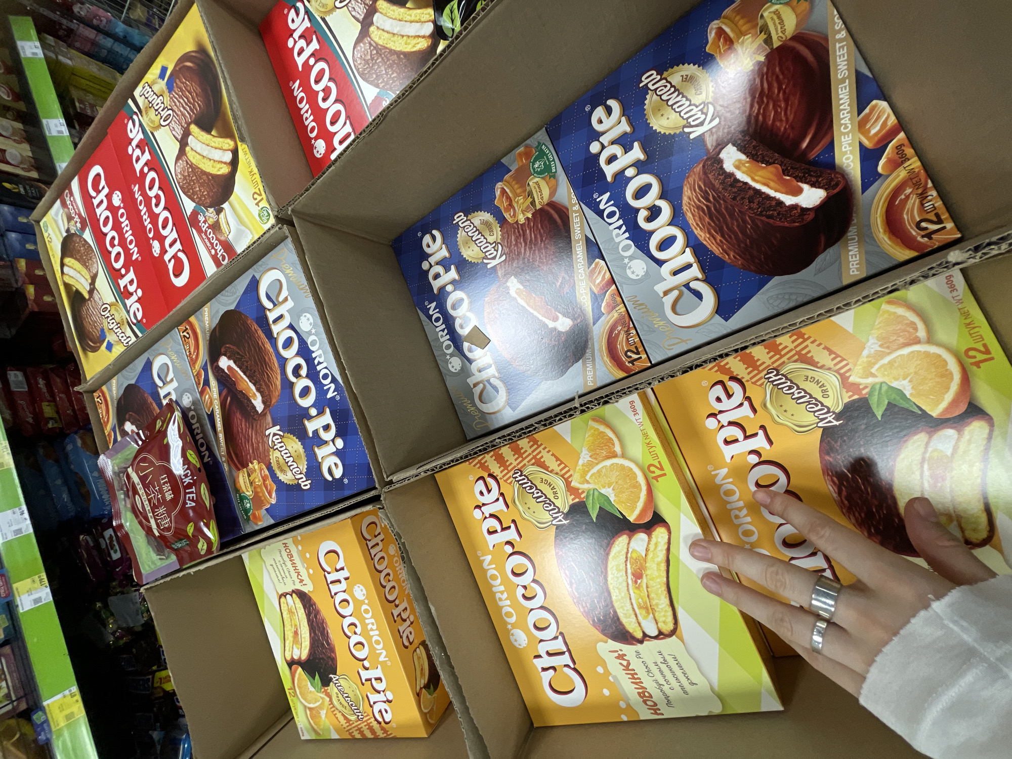This image shows a close-up view inside a cardboard box, filled with Orion Choco-Pie boxes of various flavors. Flavors visible include Original, Caramel, and Orange. There are multiple boxes of each flavor stacked inside. A hand with two silver rings is reaching towards the orange-flavored Choco-Pie boxes. The scene is likely inside a grocery store or supermarket, as evidenced by the presence of other shelves of products visible in the background. Some packaging with text in Russian is visible, and another box appears to have Chinese writing on it. There is no evidence that the location is the city of Moscow, Russia.