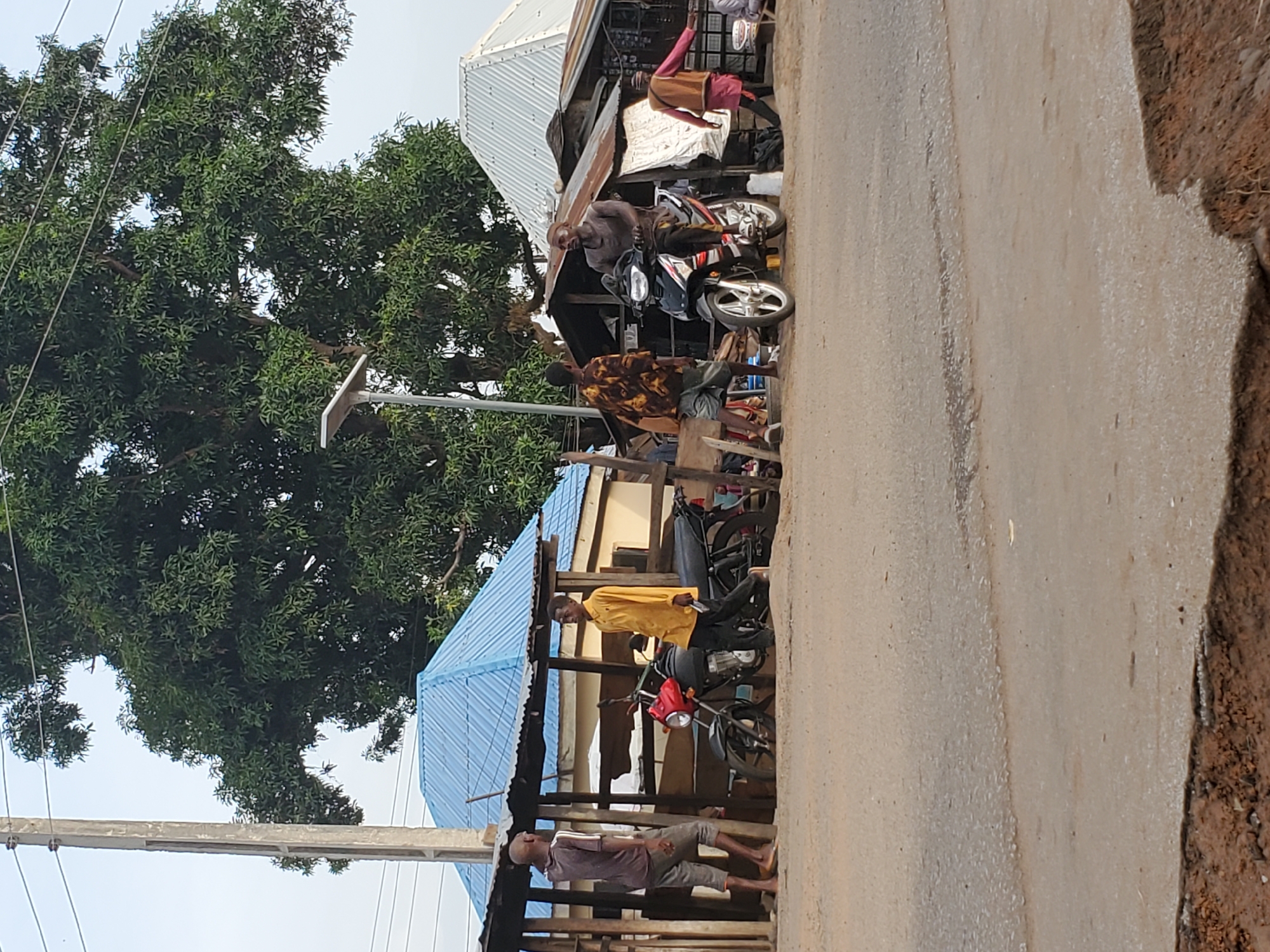 The image captures a bustling street scene in Mararaba, Nigeria, featuring multiple individuals and structures under a bright sky. A large, verdant tree with dense foliage dominates the left side of the frame, towering over the scene.
Several men are present. In the foreground, a man wearing a grey shirt and shorts stands barefoot under a wooden shelter with a blue corrugated metal roof. Nearby, another man in a yellow shirt and dark pants stands beside a red and black motorcycle, also under the blue-roofed shelter. Further back and to the right, a man in a brown top and shorts is seated on a black and silver motorcycle, positioned near another building with a white corrugated metal roof. A fourth man, dressed in a pink or reddish top, is standing near this same building, partially obscured.
The setting includes various buildings with corrugated metal roofs, suggesting a mix of residential and commercial structures, possibly small shops or market stalls. Power lines stretch across the sky, and a solar-powered street light fixture is visible near the large tree. The ground is an unpaved road, appearing as dirt or worn asphalt with some lighter streaks, possibly from water runoff. The overall atmosphere appears to be a typical daytime street activity in a lively community. No discernible text is visible in the image.