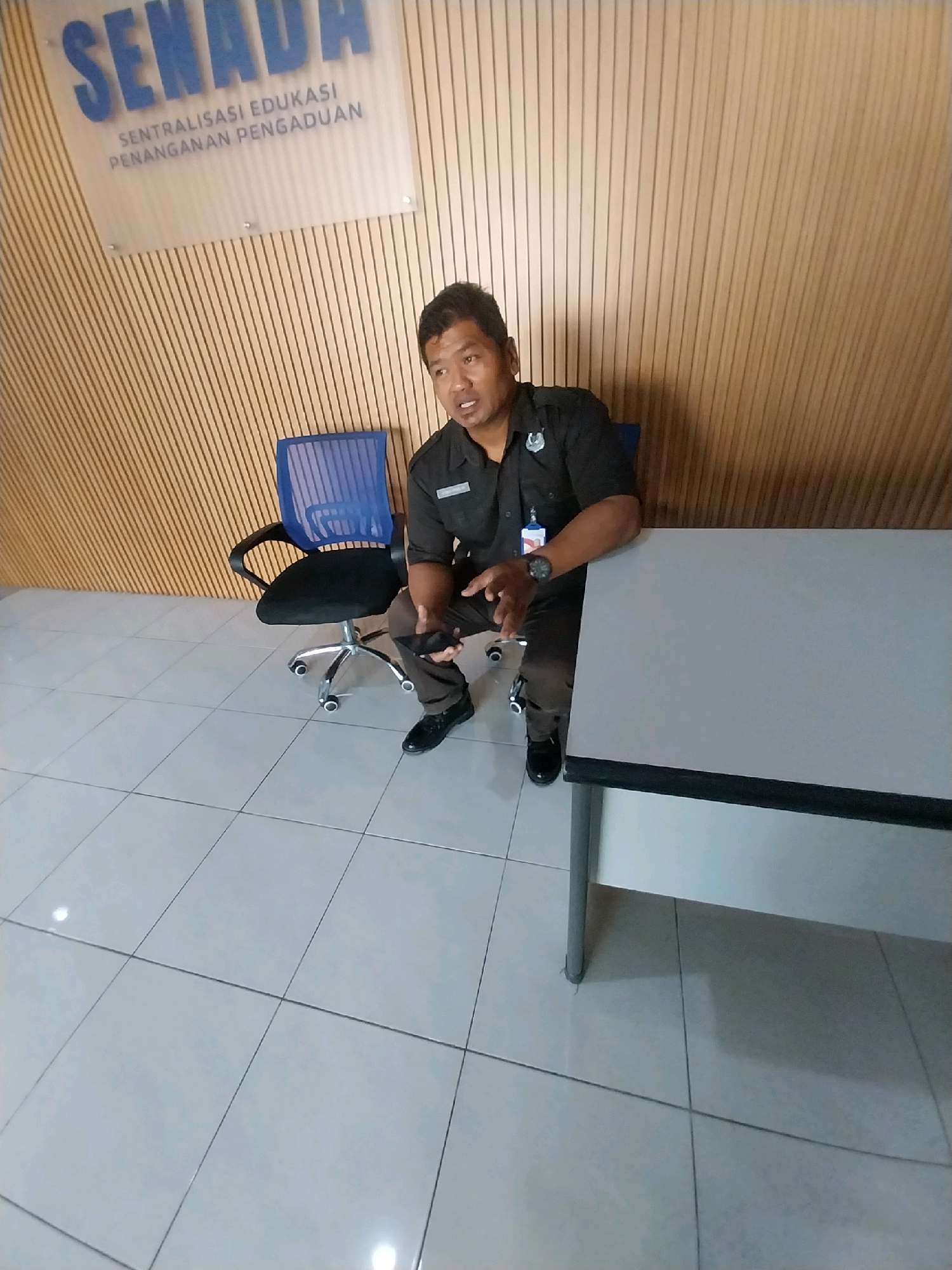 A man in a dark uniform with a badge and a lanyard is sitting on a desk in a building with wooden slatted walls and a tiled floor. He is holding a smartphone in his right hand and looking towards the camera. There is a blue mesh office chair to his left. A sign above him reads "SENADA SENTRALISASI EDUKASI PENANGANAN PENGADUAN." This sign indicates the location is likely an administrative office within the city of Depok, Indonesia, related to education and complaint handling. The man appears to be an employee or official at this location.