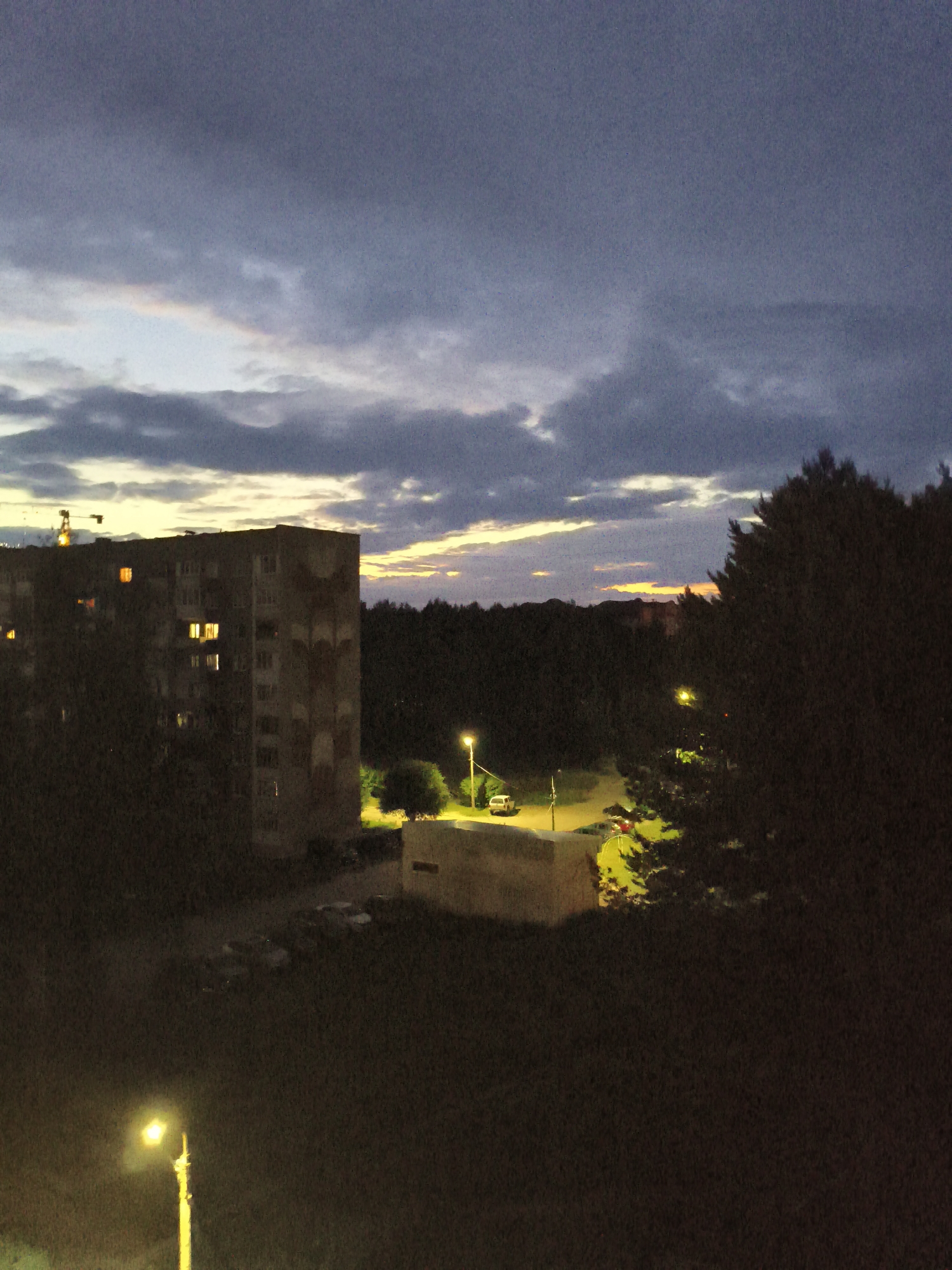 The image captures an evening scene in Chaikovsky, Russia, under a cloudy sky with hints of sunset colors. A tall, multi-story apartment building, appearing somewhat weathered, dominates the left side of the frame. Several windows on the building are illuminated, suggesting occupancy. To the right of the building, a dense line of dark trees stretches across the background. In the mid-ground, a low, rectangular structure with a lighter colored roof is visible, illuminated by a nearby streetlamp. A white car is parked to the left of this structure, and another vehicle, possibly red, is parked further to the right, partially obscured by trees. Another streetlamp stands on the right side of the frame, casting a warm glow on the surroundings. The foreground is dark and appears to be a grassy area, with a single, bright streetlamp on the far left. A construction crane is visible in the distance, above and to the left of the apartment building. The overall atmosphere is twilight, with artificial lights beginning to punctuate the dim natural light.
